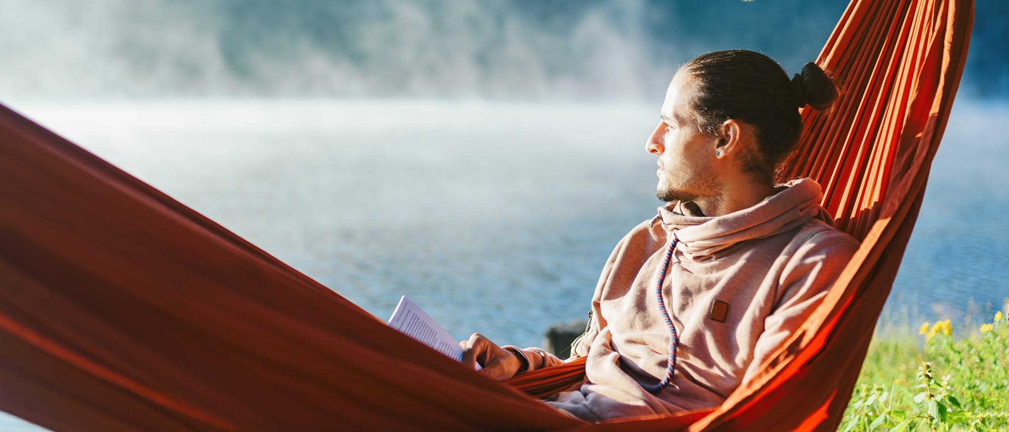 Eine Person entspannt in einer roten H&auml;ngematte am Ufer eines Sees. Sie h&auml;lt ein Buch in der Hand und blickt nachdenklich in die Ferne. Im Hintergrund ist der See mit leichtem Nebel zu sehen, umgeben von gr&uuml;ner Vegetation. Die Szene vermittelt Ruhe und Gelassenheit.