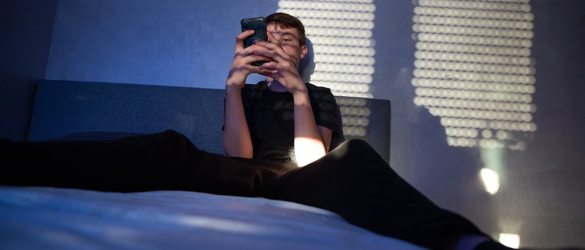 Eine junge Person sitzt auf einem Bett und h&auml;lt ein Smartphone in den H&auml;nden. Das Zimmer ist in blaues Licht getaucht, und durch ein Fenster fallen Schattenmuster an die Wand. Die Person guckt konzentriert auf das Smartphone.