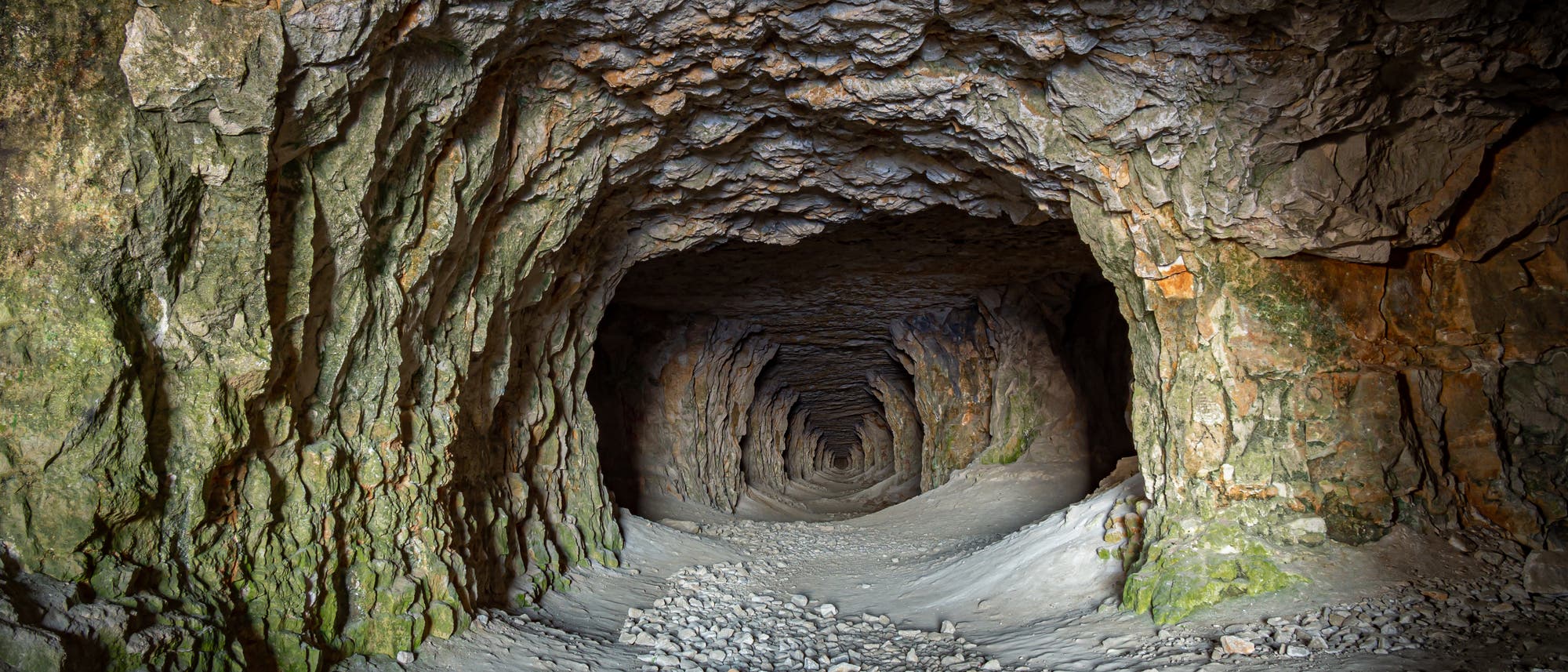 Ein langer, natürlicher Höhlentunnel mit unebenen, felsigen Wänden und einer steinigen, sandbedeckten Bodenfläche. Der Tunnel verjüngt sich in der Ferne und ist von weichem Licht erleuchtet, das die Textur der Felsen hervorhebt. Die Wände zeigen grünliche und bräunliche Farbtöne, die auf mineralische Ablagerungen hinweisen.