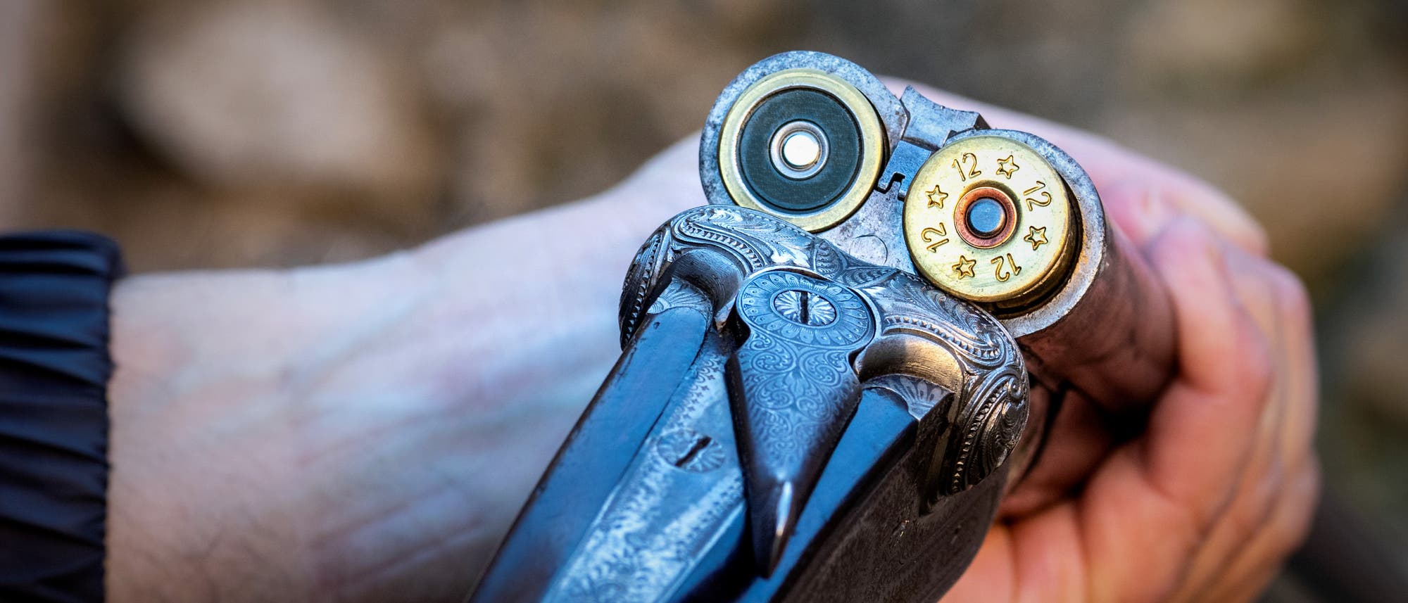 Eine Person hält eine doppelläufige Schrotflinte, die geöffnet ist, um zwei Patronen zu zeigen. Die Patronen sind mit "12" und Sternen markiert. Die Waffe hat ein kunstvolles Gravurmuster. Der Hintergrund ist unscharf und zeigt eine natürliche Umgebung. Eine Person hält eine doppelläufige Schrotflinte, die geöffnet ist, um zwei Patronen zu zeigen. Die Patronen sind mit "12" und Sternen markiert. Die Waffe hat ein kunstvolles Gravurmuster. Der Hintergrund ist unscharf und zeigt eine natürliche Umgebung.