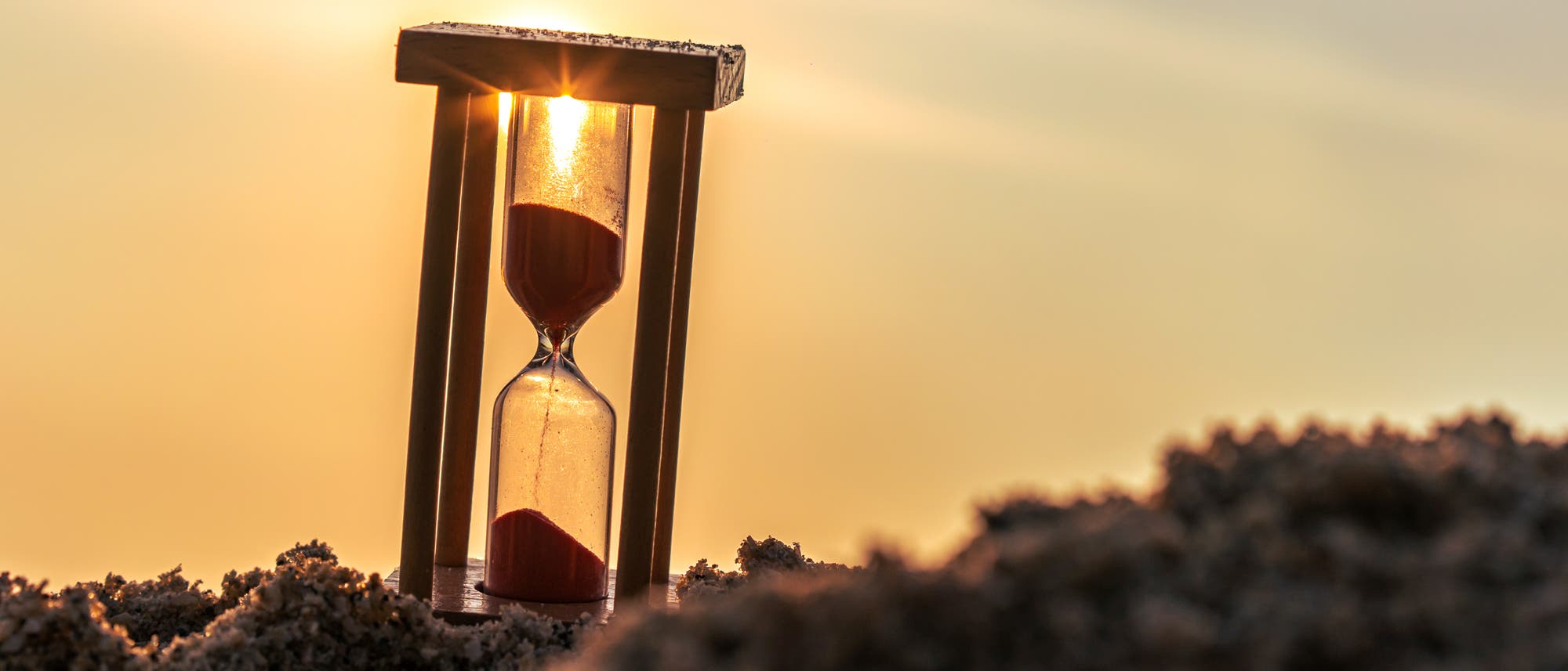 Eine Sanduhr mit rotem Sand steht auf einem sandigen Untergrund, w&auml;hrend die Sonne im Hintergrund untergeht. Der Sand rieselt langsam von der oberen in die untere Kammer, was den Fluss der Zeit symbolisiert. Die warme Beleuchtung des Sonnenuntergangs erzeugt eine ruhige und nachdenkliche Atmosph&auml;re.