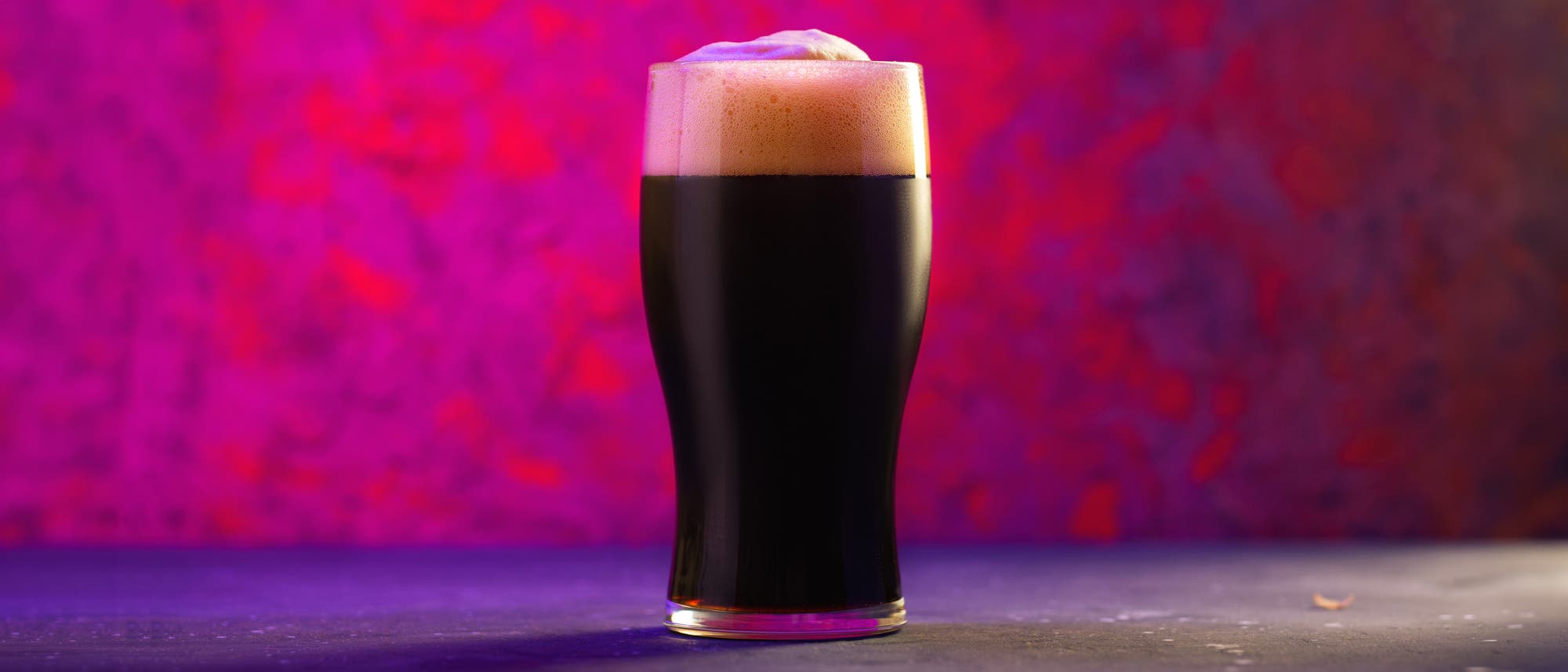 Ein Glas mit dunklem Bier, das auf einem Tisch steht. Der Schaum des Bieres ist cremig und leicht &uuml;ber den Rand des Glases gew&ouml;lbt. Der Hintergrund ist in lebhaften Rosa- und Rott&ouml;nen gehalten, was dem Bild eine stimmungsvolle Atmosph&auml;re verleiht.