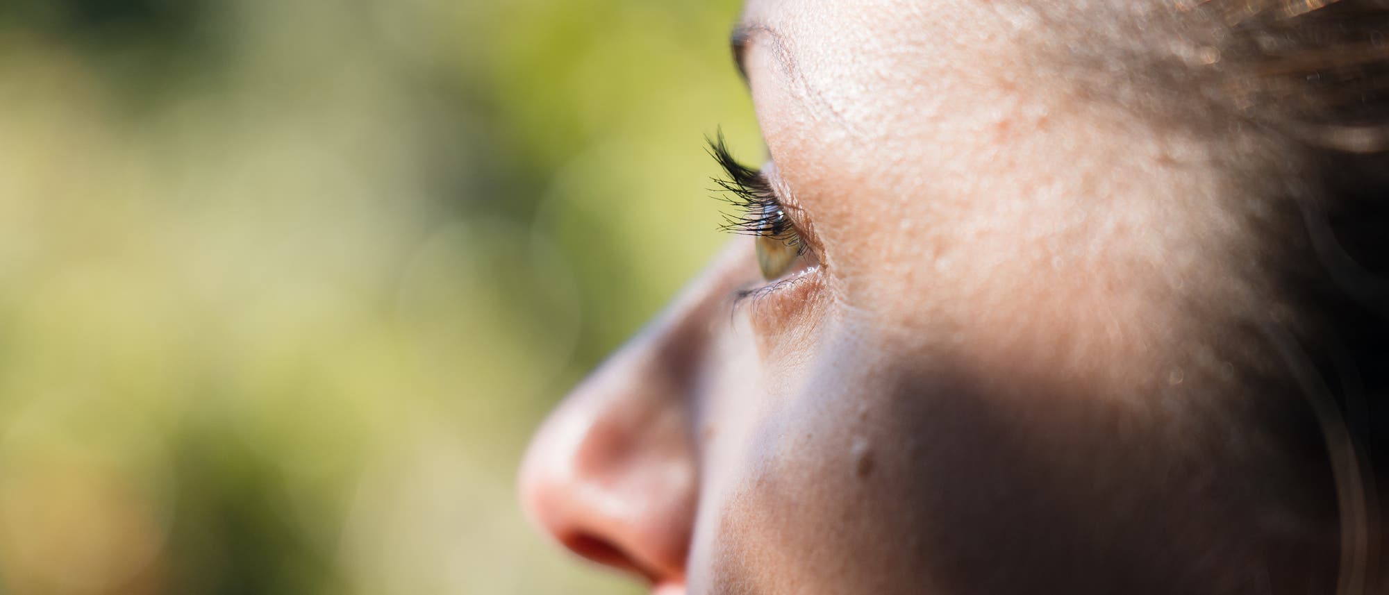 Eine Nahaufnahme eines menschlichen Gesichts von der Seite, die den Fokus auf das Auge und die Wimpern legt. Der Hintergrund ist unscharf und zeigt gr&uuml;ne, verschwommene Formen, die auf eine nat&uuml;rliche Umgebung hindeuten. Das Bild vermittelt einen ruhigen und nachdenklichen Ausdruck.