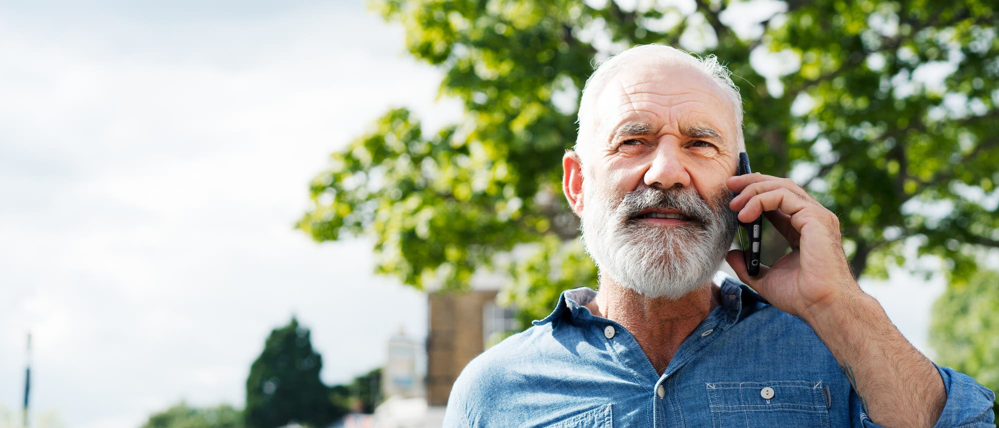 Ein älterer Mann steht im Freien und telefoniert mit einem Mobiltelefon. Er trägt ein blaues Hemd. Im Hintergrund sind Bäume und unscharfe Gebäude zu sehen, darunter eine britische Flagge. Der Himmel ist teilweise bewölkt.