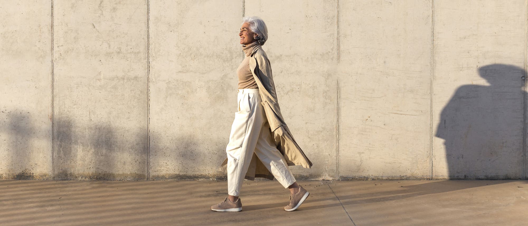 Eine Frau geht lächelnd an einer Betonwand entlang. Sie trägt einen beigen Mantel, ein helles Oberteil und weiße Hosen. Die Sonne wirft einen langen Schatten auf den Boden. Eine Frau geht lächelnd an einer Betonwand entlang. Sie trägt einen beigen Mantel, ein helles Oberteil und weiße Hosen. Die Sonne wirft einen langen Schatten auf den Boden.