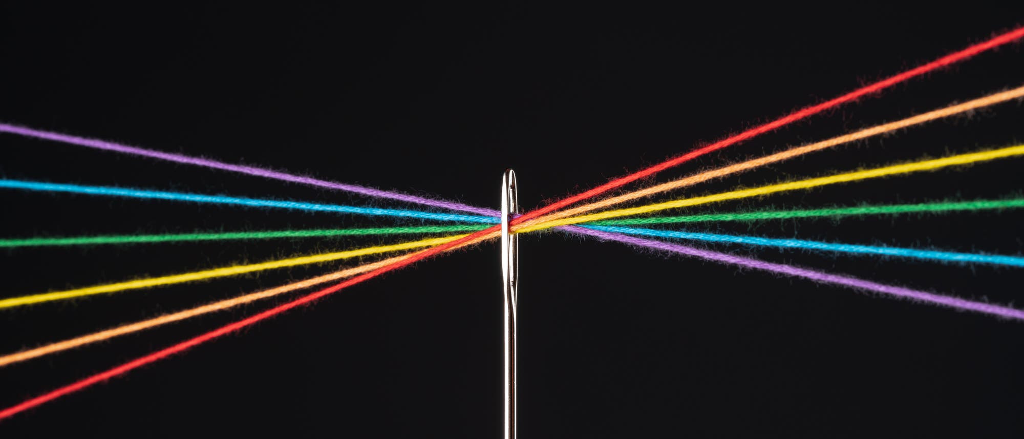 Eine Nähnadel mit sechs bunten Fäden, die durch das Nadelöhr gezogen sind. Die Fäden haben die Farben Rot, Orange, Gelb, Grün, Blau und Lila und sind zu beiden Seiten der Nadel gespannt. Der Hintergrund ist schwarz, was die farbigen Fäden hervorhebt. Eine Nähnadel mit sechs bunten Fäden, die durch das Nadelöhr gezogen sind. Die Fäden haben die Farben Rot, Orange, Gelb, Grün, Blau und Lila und sind zu beiden Seiten der Nadel gespannt. Der Hintergrund ist schwarz, was die farbigen Fäden hervorhebt.