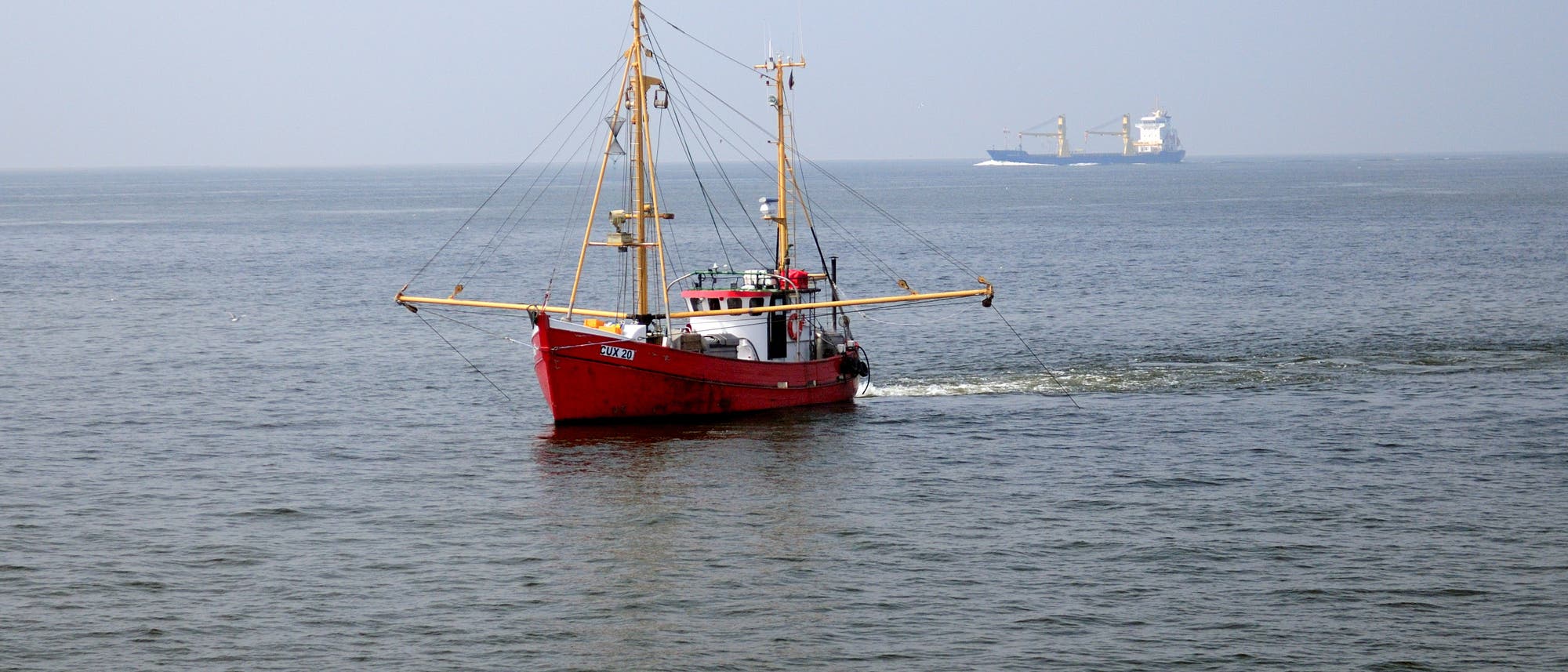 ein Foto zeigt einen roten Trawler mit ausgefahrenen Grundschleppnetzen in der Nordsee, im Hintergrund ist ein Frachter zu sehen