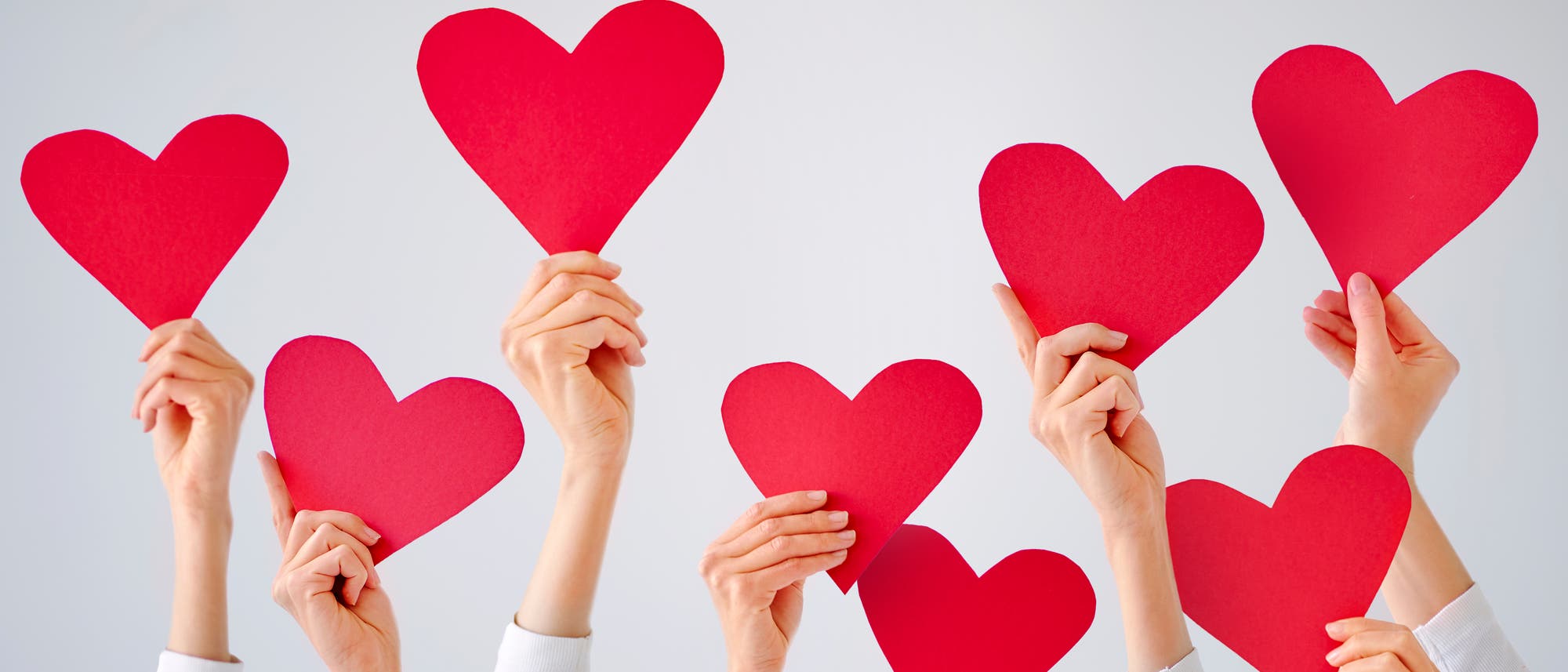 Mehrere H&auml;nde halten rote Papierherzen vor einem hellen Hintergrund hoch. Die Szene vermittelt ein Gef&uuml;hl von Gemeinschaft und Unterst&uuml;tzung.