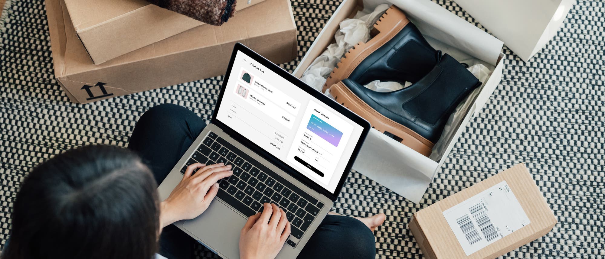 Eine Person sitzt auf einem Teppichboden und benutzt einen Laptop, um online einzukaufen. Auf dem Bildschirm ist eine Checkout-Seite zu sehen. Um die Person herum liegen geöffnete Pakete mit Schuhen und einem gefalteten Schal.