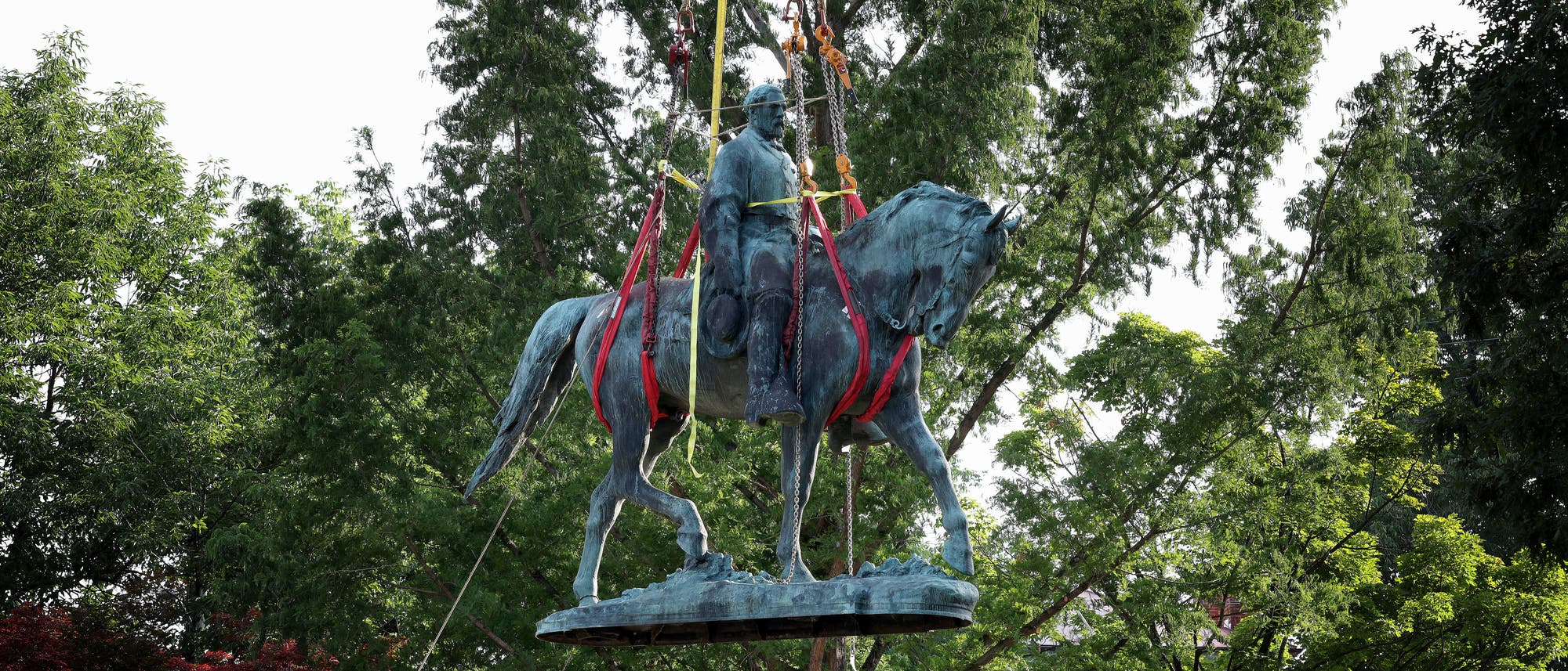 Eine massive Reiterstatue schwebt, von starken Gurten gehalten, durch die Luft. Sie ist von grünen Bäumen umgeben. Eine massive Reiterstatue schwebt, von starken Gurten gehalten, durch die Luft. Sie ist von grünen Bäumen umgeben.