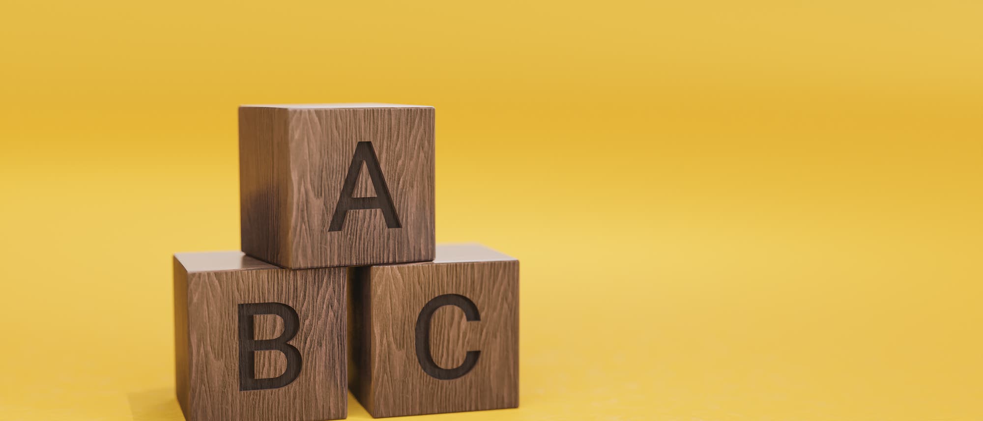 Drei hölzerne Würfel mit den Buchstaben A, B und C sind auf einem gelben Hintergrund gestapelt. Der Würfel mit dem Buchstaben A befindet sich oben, während B und C unten nebeneinander stehen.