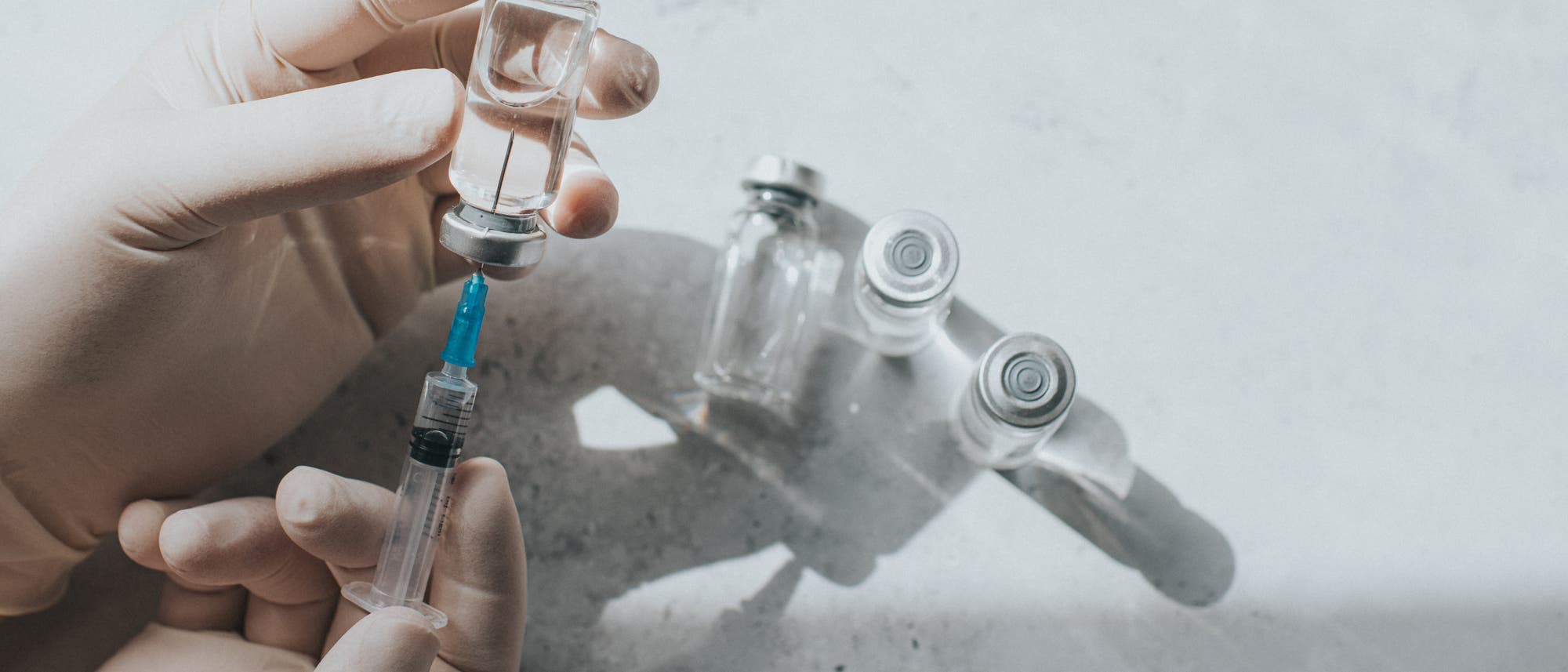 Eine Person mit medizinischen Handschuhen zieht Flüssigkeit aus einem Glasfläschchen in eine Spritze auf. Im Hintergrund sind weitere leere Fläschchen auf einer hellen Oberfläche zu sehen. Die Szene ist gut beleuchtet und vermittelt einen klinischen, sterilen Eindruck.