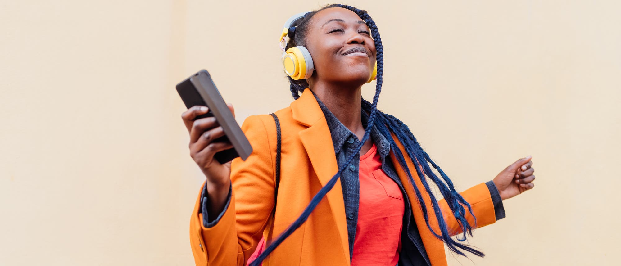 Eine Person steht vor einer beigen Wand, tr&auml;gt gelbe Kopfh&ouml;rer und h&auml;lt ein Smartphone in der Hand. Sie tr&auml;gt einen orangefarbenen Blazer, ein rotes Oberteil und blaue Jeans. Die Person wirkt entspannt und genie&szlig;t die Musik.
