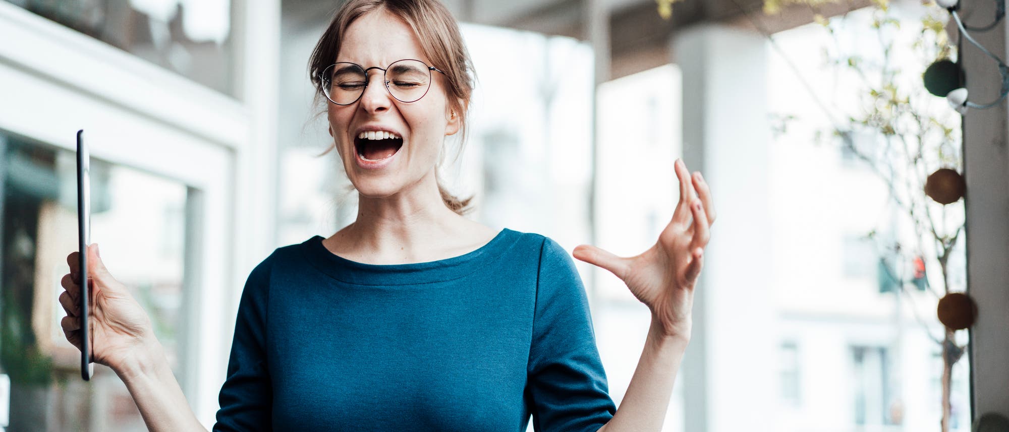 Eine junge Frau in einem blauen Oberteil steht in einem modernen Büro. Sie hält beide Arme leicht erhoben, in einer Hand ein Tablet. Ihre Mimik wirkt gequält und angestrengt: Sie hat die Augen zusammengekniffen und den Mund weit offen, als würde sie schreien. Eine junge Frau in einem blauen Oberteil steht in einem modernen Büro. Sie hält beide Arme leicht erhoben, in einer Hand ein Tablet. Ihre Mimik wirkt gequält und angestrengt: Sie hat die Augen zusammengekniffen und den Mund weit offen, als würde sie schreien.
