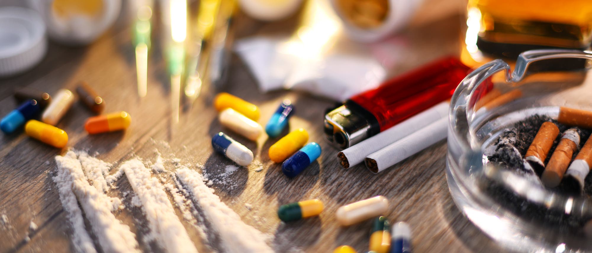 Eine Nahaufnahme eines Tisches mit verschiedenen Drogen und Utensilien. Zu sehen sind bunte Pillen, weiße Pulverlinien, Spritzen, Zigarettenstummel in einem Aschenbecher, ein Feuerzeug und ein Glas mit einer bernsteinfarbenen Flüssigkeit. Die Szene vermittelt ein Bild von Substanzmissbrauch und Sucht.