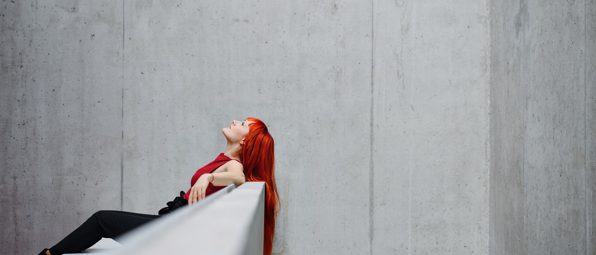 Eine Person mit leuchtend rotem Haar lehnt entspannt an einer Betonwand. Sie trägt ein rotes Oberteil und schwarze Hosen. Der Blick ist nach oben gerichtet, als ob sie den Himmel betrachtet. Die Umgebung ist minimalistisch und modern, mit glatten, grauen Betonflächen. Eine Person mit leuchtend rotem Haar lehnt entspannt an einer Betonwand. Sie trägt ein rotes Oberteil und schwarze Hosen. Der Blick ist nach oben gerichtet, als ob sie den Himmel betrachtet. Die Umgebung ist minimalistisch und modern, mit glatten, grauen Betonflächen.