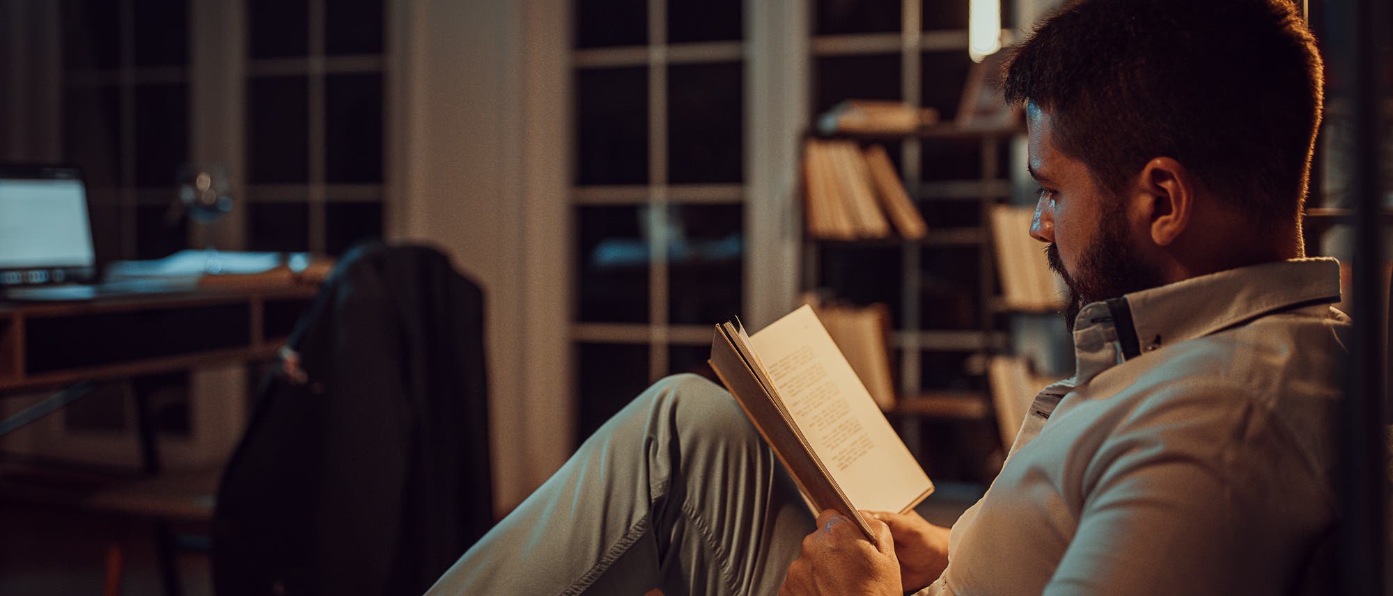 Ein Mann sitzt in einem gemütlichen Raum und liest ein Buch. Er trägt ein helles Hemd und eine Uhr am Handgelenk. Im Hintergrund sind ein Schreibtisch mit einem Laptop und ein Bücherregal zu sehen. Der Raum ist schwach beleuchtet, was eine entspannte Atmosphäre schafft.