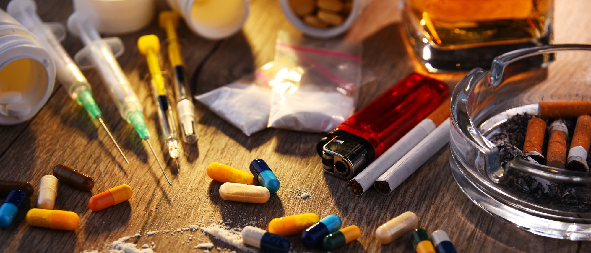 Eine Nahaufnahme eines Tisches mit verschiedenen Drogen und Utensilien. Zu sehen sind bunte Pillen, weiße Pulverlinien, Spritzen, Zigarettenstummel in einem Aschenbecher, ein Feuerzeug und ein Glas mit einer bernsteinfarbenen Flüssigkeit. Die Szene vermittelt ein Bild von Substanzmissbrauch und Sucht.