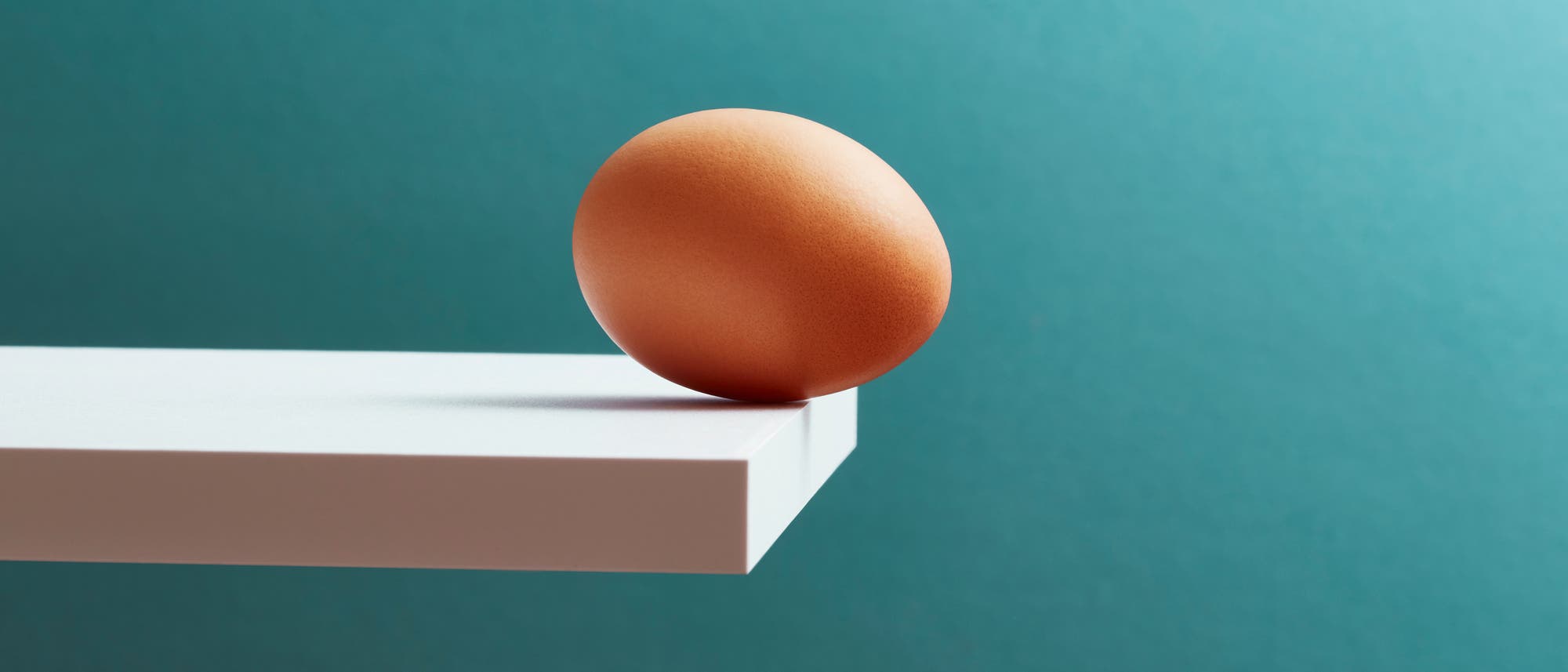 Ein braunes Ei balanciert am Rand einer weißen, wie ein Sprungbrett wirkenden Plattform vor einem türkisfarbenen Hintergrund. Das Bild vermittelt ein Gefühl von Spannung und Balance. Es gibt keinen Text oder eine URL im Bild. Ein braunes Ei balanciert am Rand einer weißen, wie ein Sprungbrett wirkenden Plattform vor einem türkisfarbenen Hintergrund. Das Bild vermittelt ein Gefühl von Spannung und Balance. Es gibt keinen Text oder eine URL im Bild.