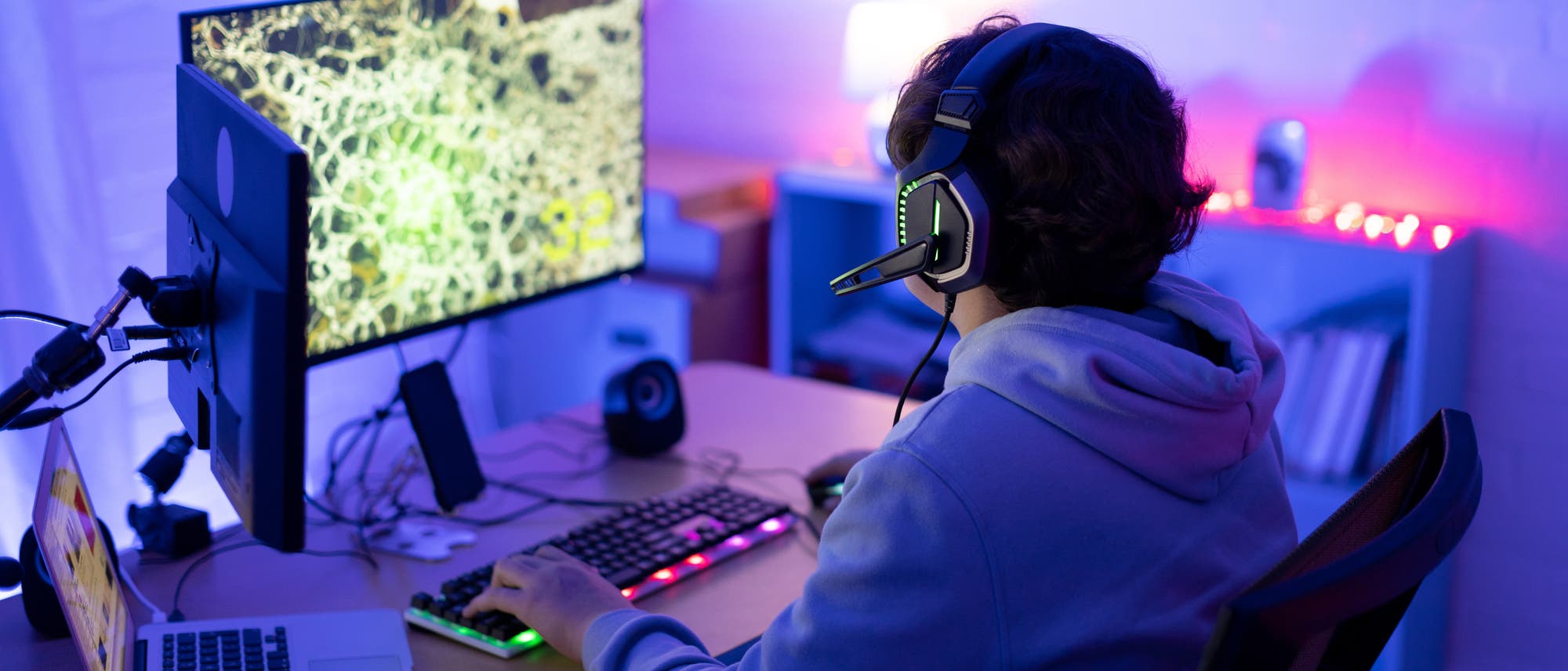 Eine Person sitzt an einem Schreibtisch und spielt ein Computerspiel. Sie trägt ein Headset und schaut auf einen großen Monitor, der ein Spiel mit einer detaillierten Karte zeigt. Auf dem Schreibtisch befinden sich eine beleuchtete Tastatur, eine Maus, ein Laptop und ein Mikrofon. Der Raum ist in blauem und violettem Licht beleuchtet, was eine Gaming-Atmosphäre schafft. Im Hintergrund sind Regale mit Büchern und Dekorationen zu sehen.