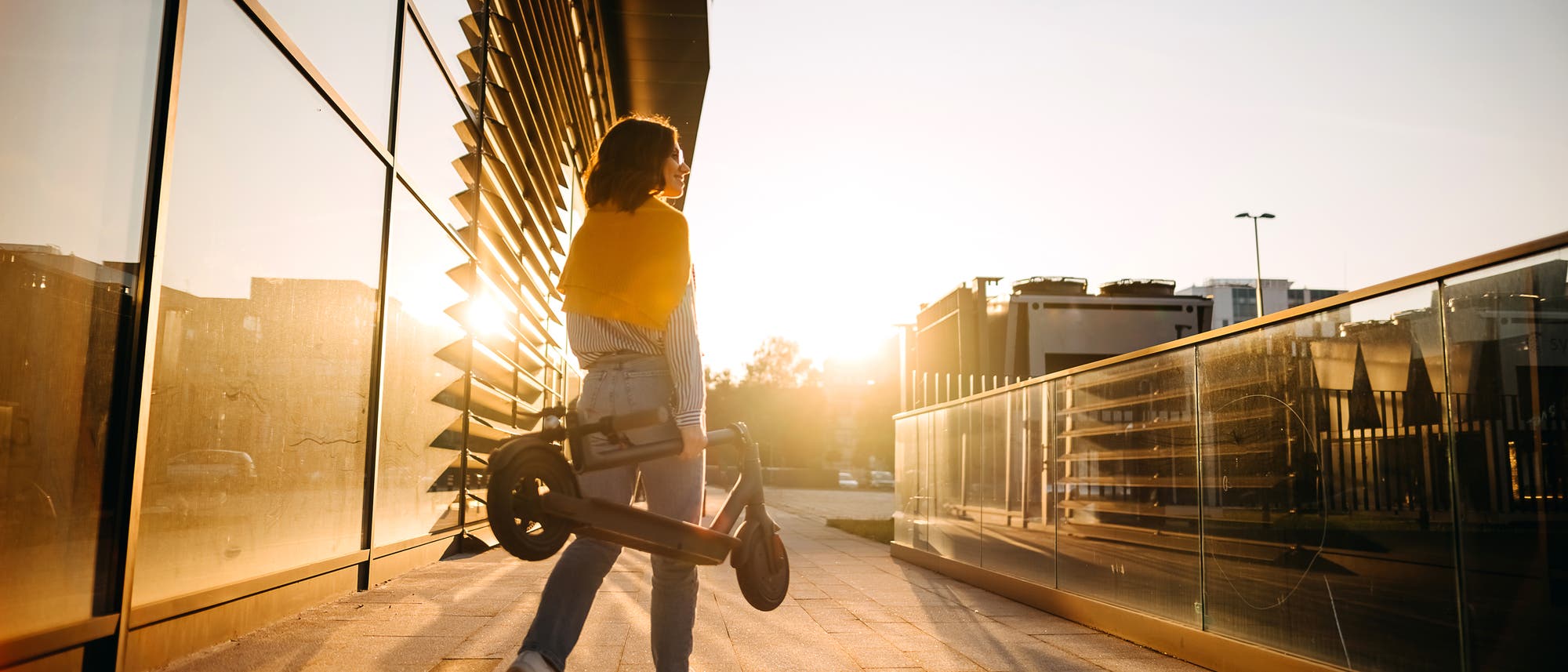 Eine Person geht bei Sonnenuntergang entlang eines modernen Gebäudes mit Glasfassade. Sie trägt einen zusammengeklappten Elektroroller und hat eine gelbe Jacke über die Schultern gelegt. Die Szene ist in warmes, goldenes Licht getaucht, das lange Schatten auf den Boden wirft. Im Hintergrund sind städtische Strukturen und ein klarer Himmel zu sehen.