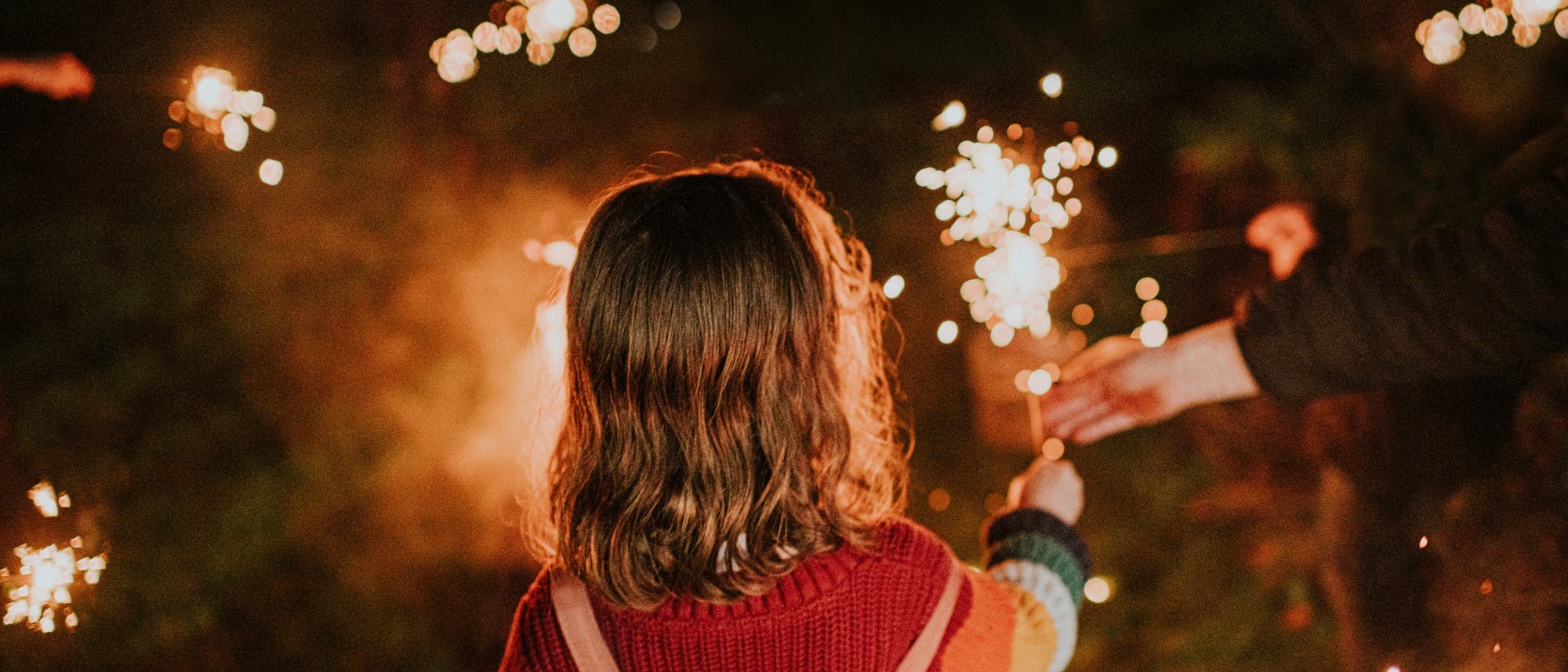 Ein Kind in einem gestreiften Pullover und Latzhose h&auml;lt eine Wunderkerze in der Hand. Es ist Nacht, und im Hintergrund sind weitere Lichter zu sehen. Eine andere Hand reicht dem Kind eine weitere Wunderkerze. Die Szene ist in warmes Licht getaucht und vermittelt eine festliche Atmosph&auml;re.