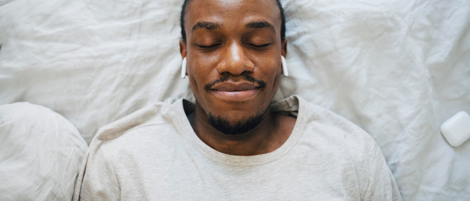 Eine Person liegt entspannt auf einem Bett mit wei&szlig;en Laken und tr&auml;gt kabellose Ohrh&ouml;rer. Die Augen sind geschlossen, und die Person wirkt zufrieden. Neben dem Kopf liegt ein Ladeetui f&uuml;r die Ohrh&ouml;rer.
