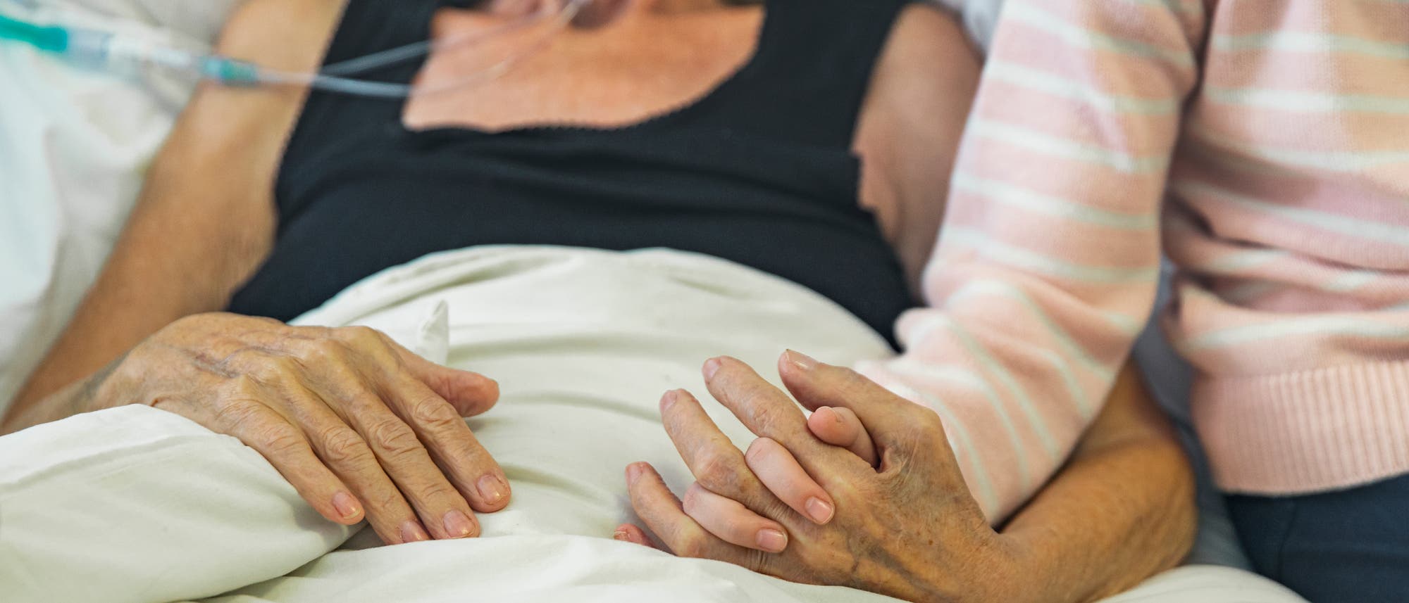 Eine ältere Frau liegt im Krankenhausbett, mit einer Sauerstoffnasenbrille verbunden. Ein Kind sitzt neben ihr und hält sanft ihre Hand. Eine ältere Frau liegt im Krankenhausbett, mit einer Sauerstoffnasenbrille verbunden. Ein Kind sitzt neben ihr und hält sanft ihre Hand.