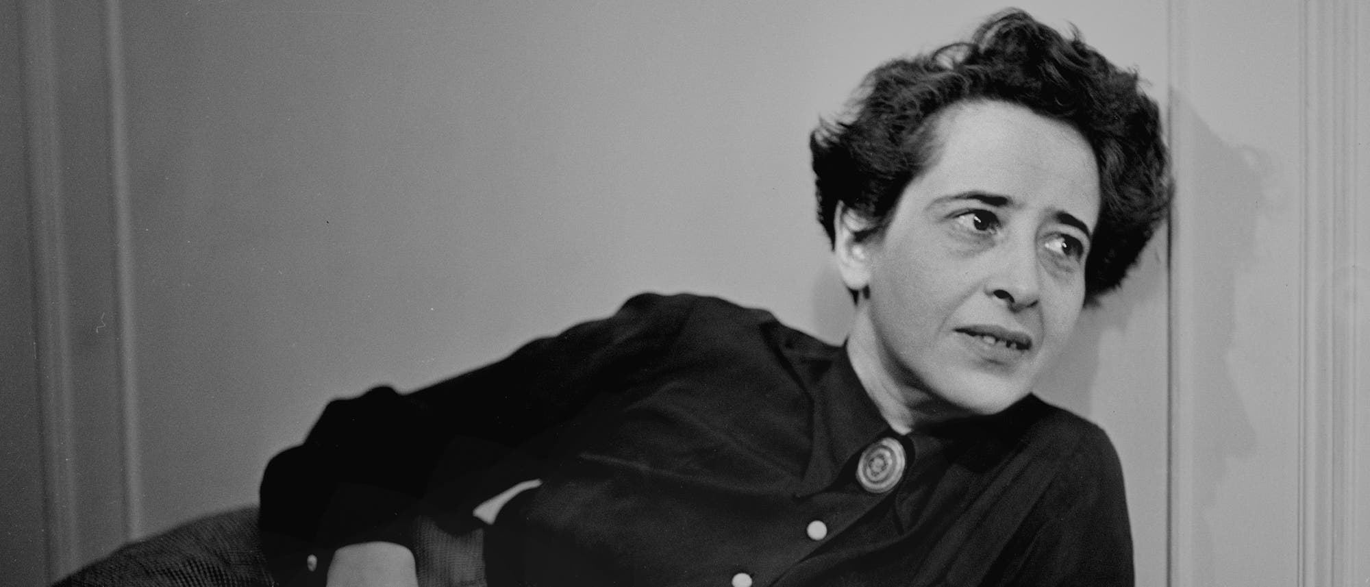 Eine Person lehnt sich an eine Wand und blickt nachdenklich zur Seite. Sie trägt ein dunkles Oberteil mit einem runden Schmuckstück am Kragen. Der Hintergrund ist schlicht und hell, was den Fokus auf die Person lenkt.