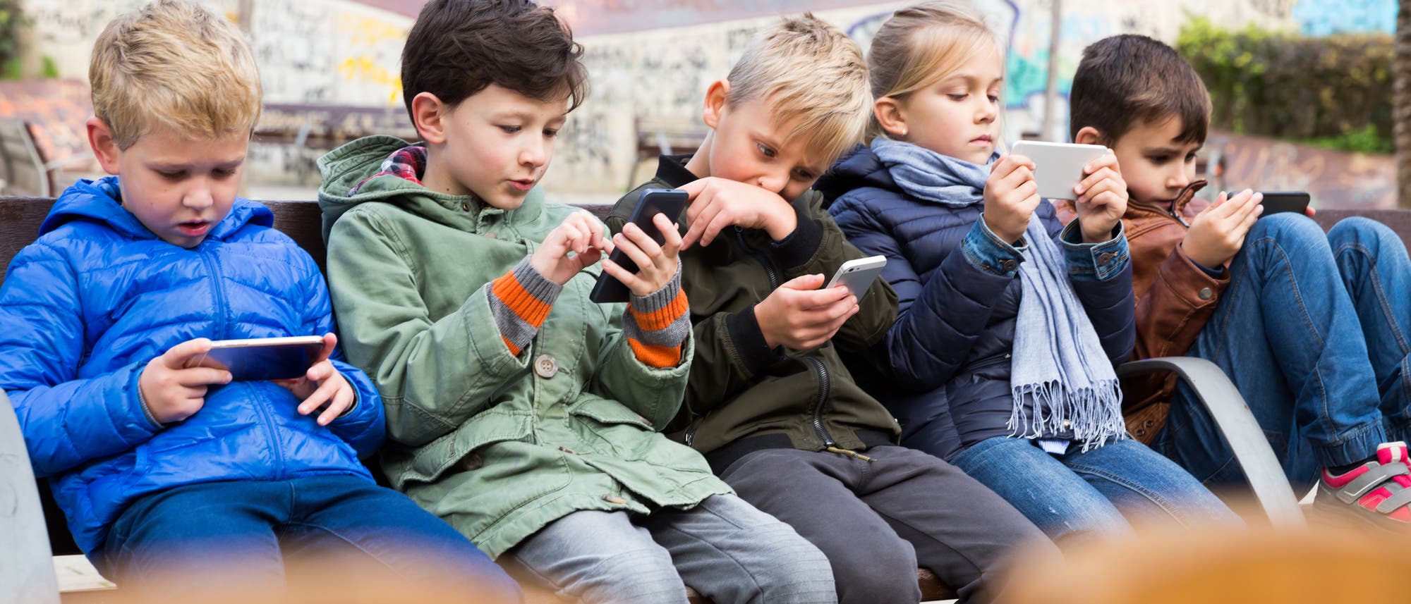 Fünf Kinder sitzen auf einer Bank im Freien und konzentrieren sich auf ihre Smartphones. Sie tragen Jacken. Im Hintergrund ist eine mit Graffiti bedeckte Wand zu sehen. Jedes Kind ist in sein Handy vertieft, was auf die Nutzung von Technologie in der Freizeit hinweist.