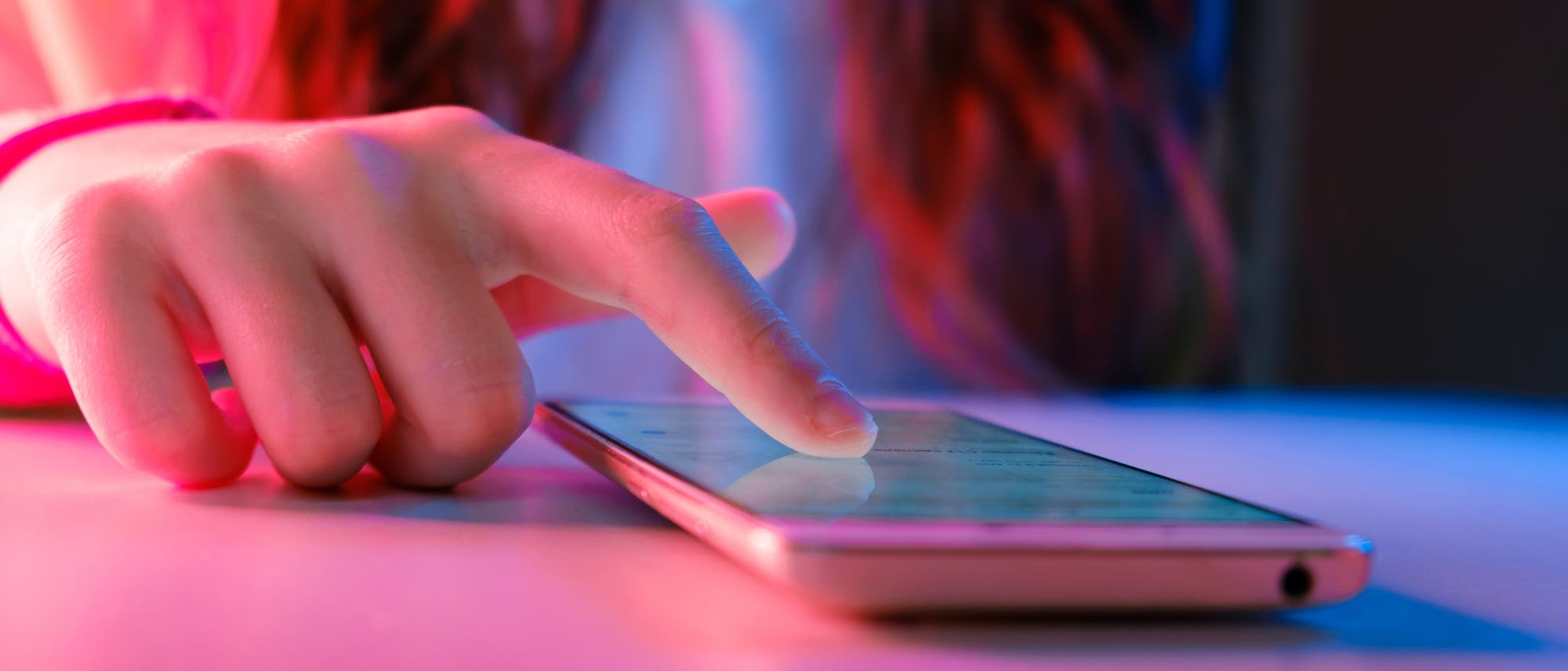 Eine Person ber&uuml;hrt mit dem Zeigefinger den Bildschirm eines Smartphones, das auf einem Tisch liegt. Die Szene ist in farbiges Licht getaucht, das einen rosa und blauen Schimmer erzeugt. Der Fokus liegt auf der Interaktion mit dem Ger&auml;t, was auf die Nutzung von Technologie hinweist.