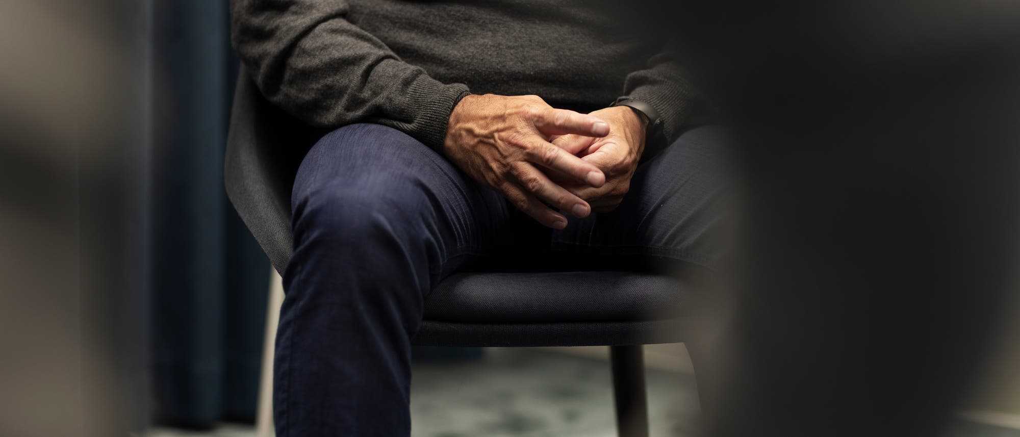 Eine Person sitzt auf einem Stuhl, die H&auml;nde sind gefaltet und ruhen auf dem Scho&szlig;. Die Person tr&auml;gt einen grauen Pullover und blaue Jeans. Der Hintergrund ist unscharf, mit einem Vorhang auf der linken Seite. Die Szene wirkt ruhig und nachdenklich.