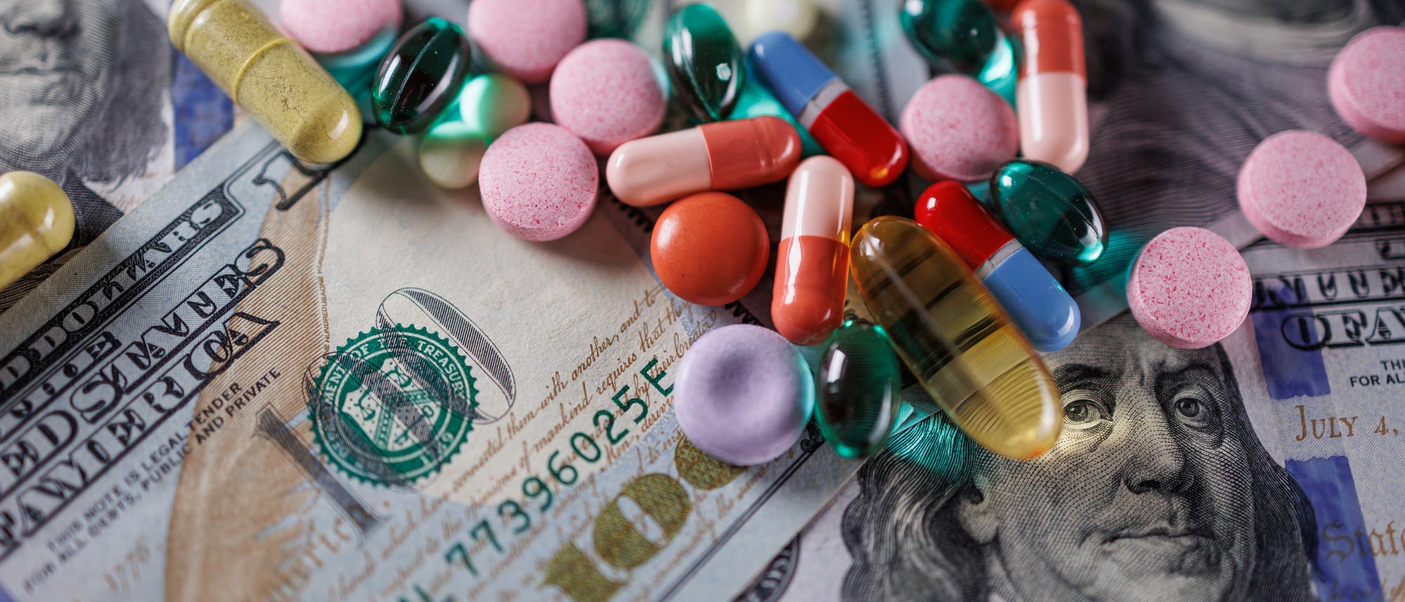 Verschiedene bunte Pillen und Kapseln liegen verstreut auf mehreren 100-Dollar-Scheinen. Die Szene symbolisiert die Verbindung zwischen Geld und der Pharmaindustrie. Im Hintergrund ist das Porträt von Benjamin Franklin auf den Geldscheinen zu sehen. Verschiedene bunte Pillen und Kapseln liegen verstreut auf mehreren 100-Dollar-Scheinen. Die Szene symbolisiert die Verbindung zwischen Geld und der Pharmaindustrie. Im Hintergrund ist das Porträt von Benjamin Franklin auf den Geldscheinen zu sehen.