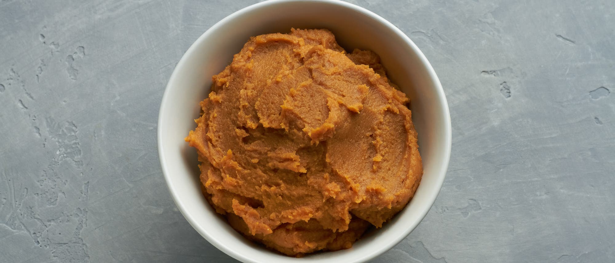 Eine weiße Schüssel mit einer Portion brauner Miso-Paste, auf einer grauen, strukturierten Oberfläche platziert. Die Paste hat eine glatte, cremige Konsistenz. Eine weiße Schüssel mit einer Portion brauner Miso-Paste, auf einer grauen, strukturierten Oberfläche platziert. Die Paste hat eine glatte, cremige Konsistenz.