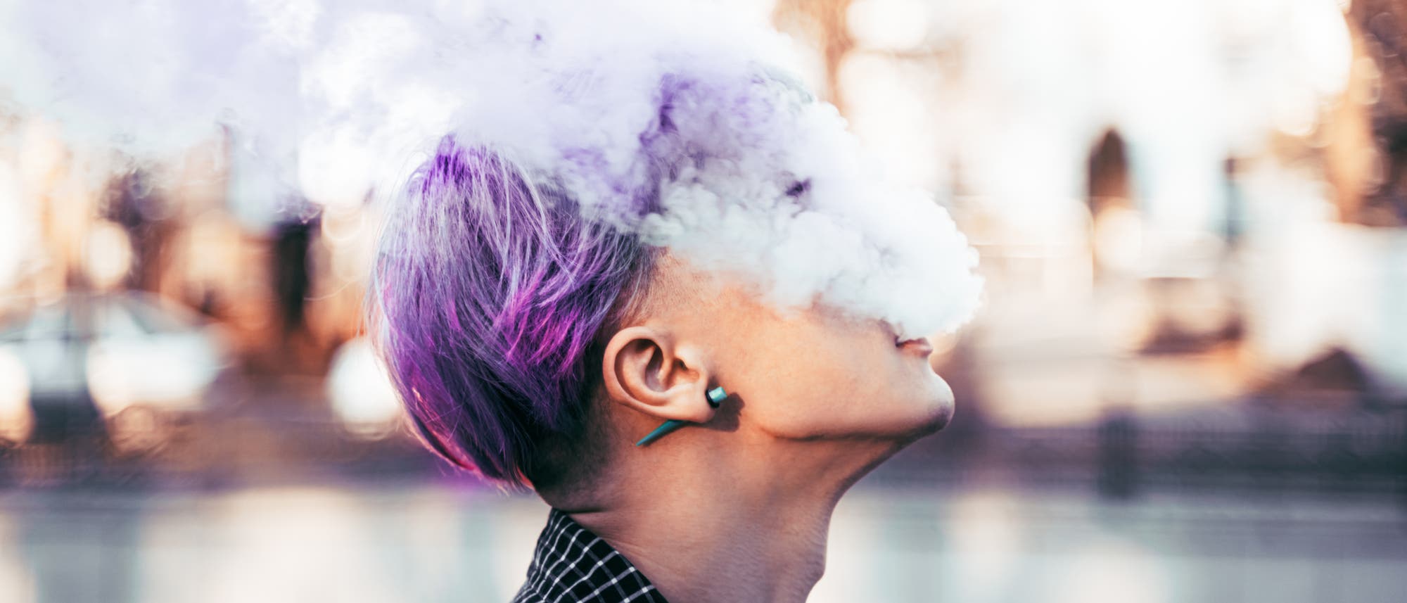 Eine Person mit lila Haaren bläst eine große Wolke aus Dampf in die Luft. Der Hintergrund ist unscharf und zeigt eine städtische Umgebung mit Bäumen und Gebäuden. Die Szene vermittelt eine entspannte Atmosphäre. Eine Person mit lila Haaren bläst eine große Wolke aus Dampf in die Luft. Der Hintergrund ist unscharf und zeigt eine städtische Umgebung mit Bäumen und Gebäuden. Die Szene vermittelt eine entspannte Atmosphäre.