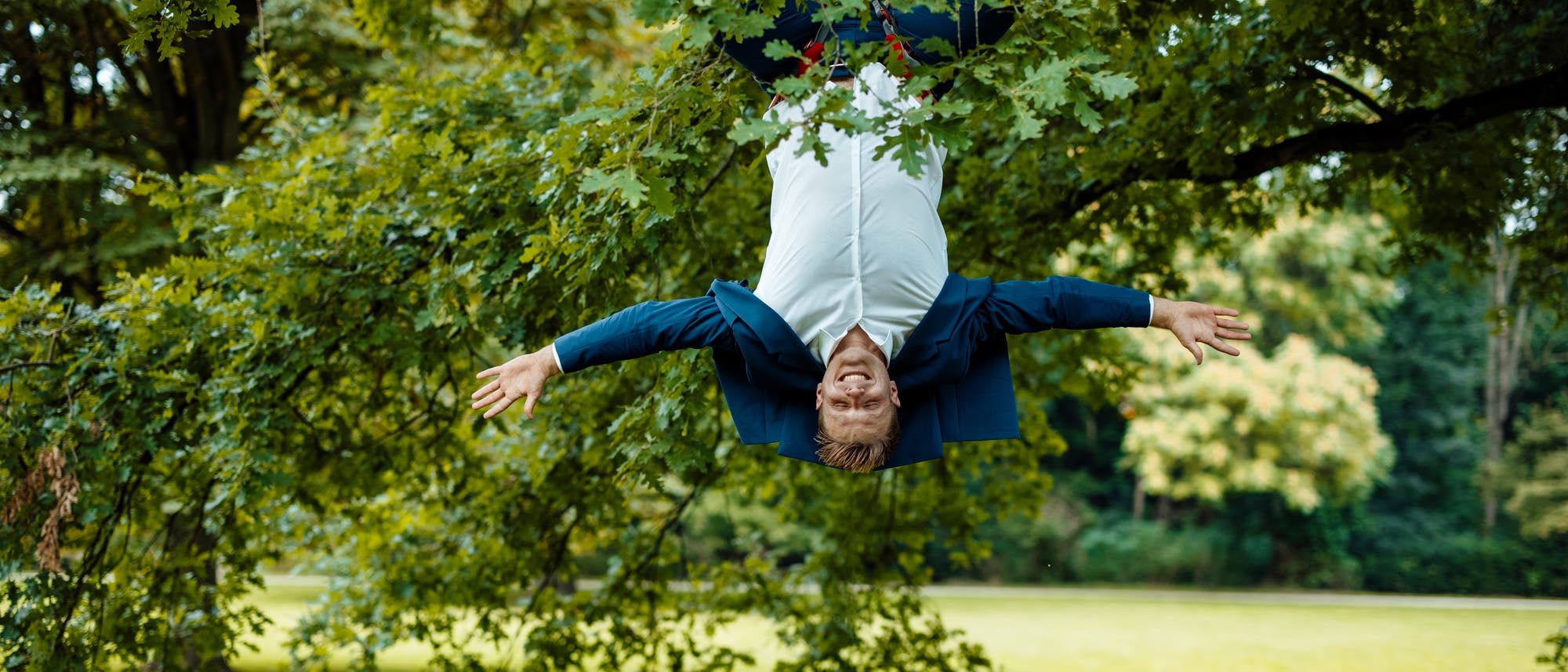 Ein Mann in weißem Hemd und einem blauen Anzug hängt kopfüber an einem Baum in einem Park. Er lächelt und breitet die Arme aus. Im Hintergrund sind grüne Wiesen und Bäume zu sehen.