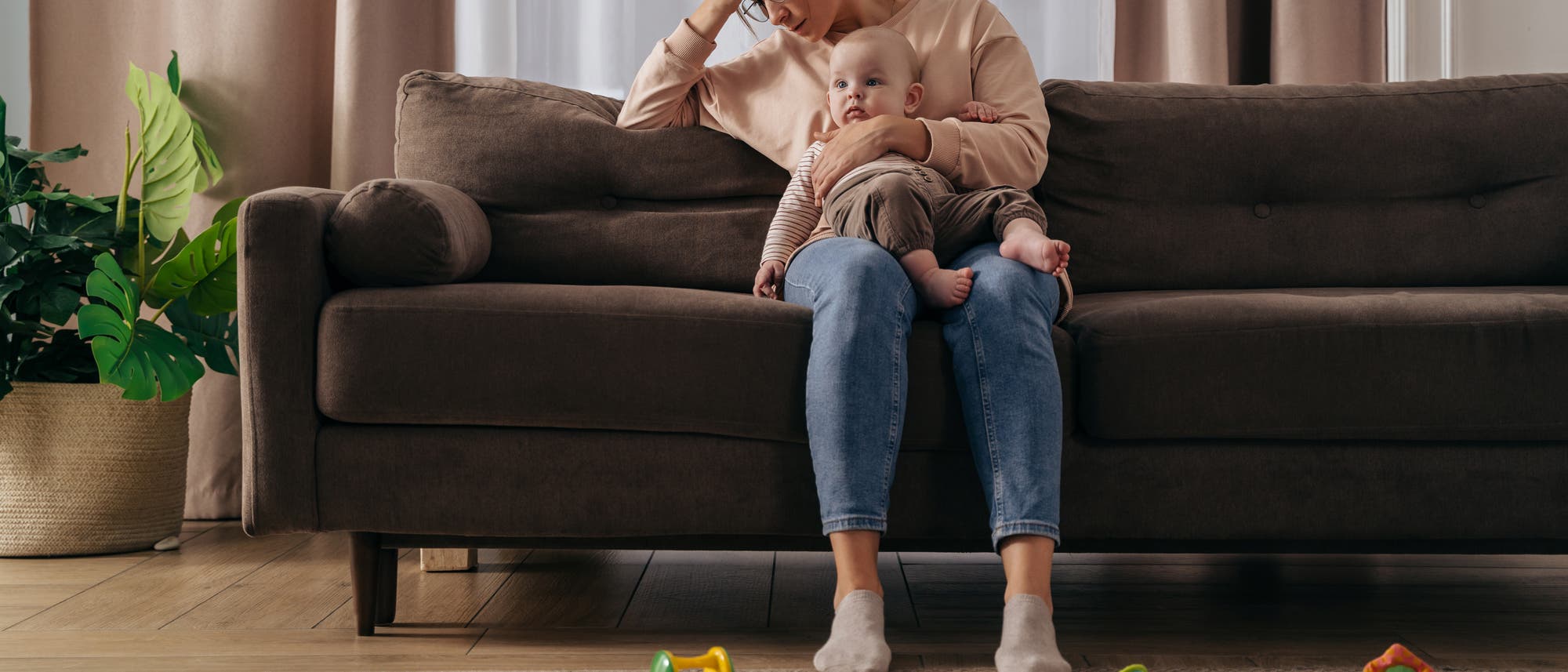 Eine Mutter sitzt auf einem Sofa und h&auml;lt ihr Baby, sie sieht m&uuml;de und gestresst aus.