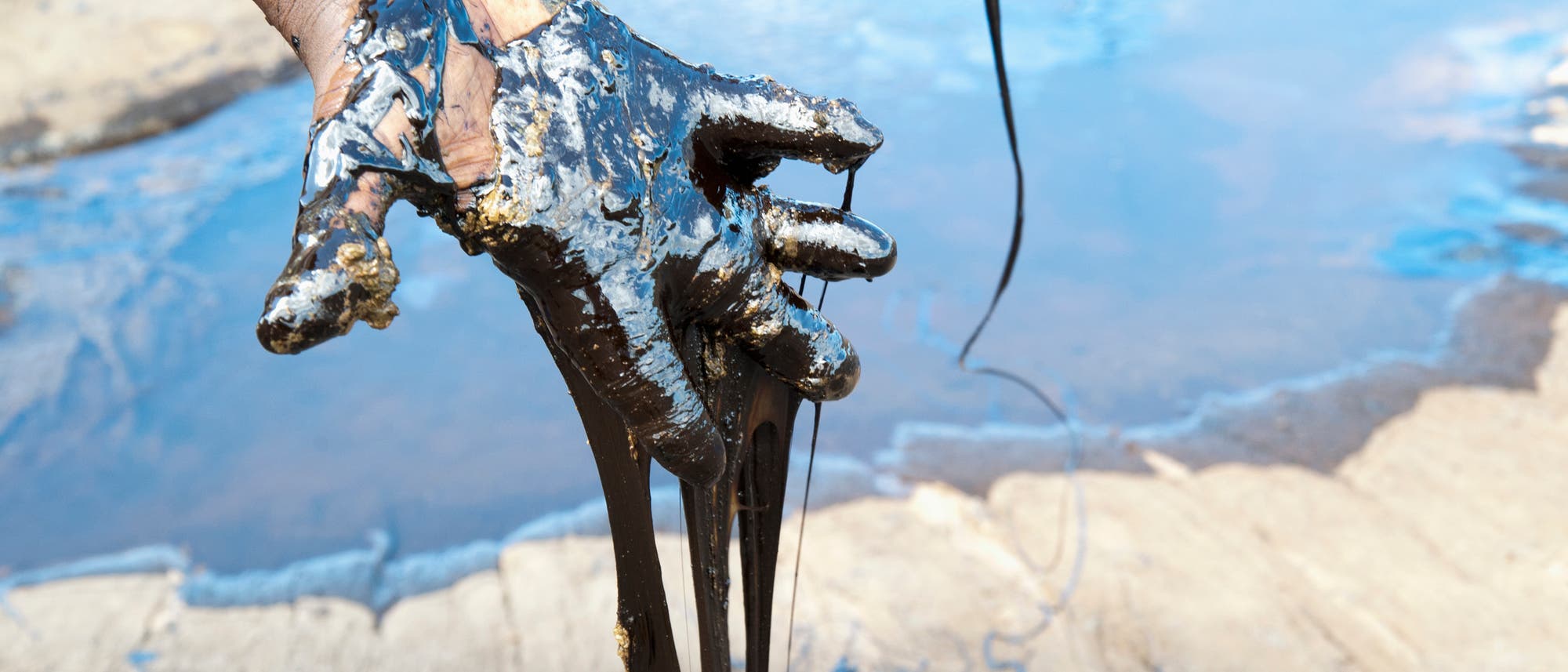 Eine Hand, die mit dickem, schwarzem &Ouml;l bedeckt ist, h&auml;lt eine z&auml;hfl&uuml;ssige Masse. Im Hintergrund ist eine sandige Oberfl&auml;che mit Pf&uuml;tzen aus &Ouml;l zu sehen. Die Szene vermittelt den Eindruck von Umweltverschmutzung und &Ouml;lversch&uuml;ttung.