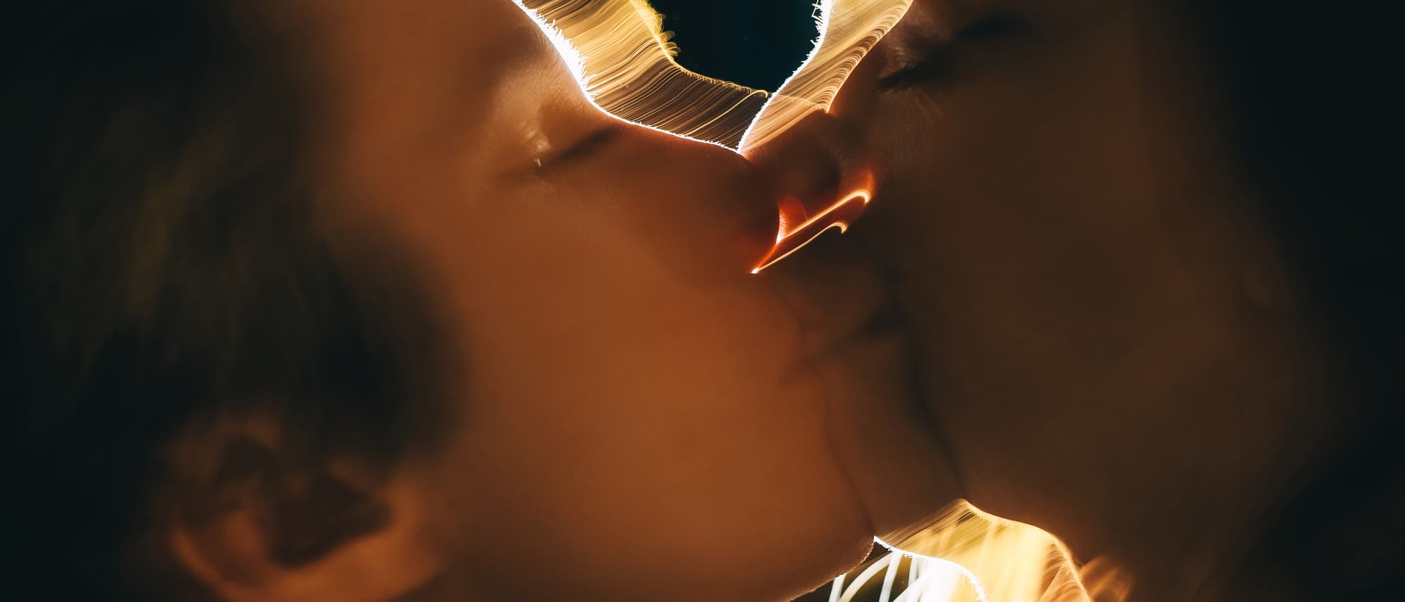 Ein k&uuml;nstlerisches Foto von zwei Personen, die sich k&uuml;ssen. Die Szene ist in warmes, goldenes Licht getaucht, das die Konturen der Gesichter betont und eine romantische Atmosph&auml;re schafft. Die Beleuchtung erzeugt weiche, flie&szlig;ende Linien um die Silhouetten der Personen.