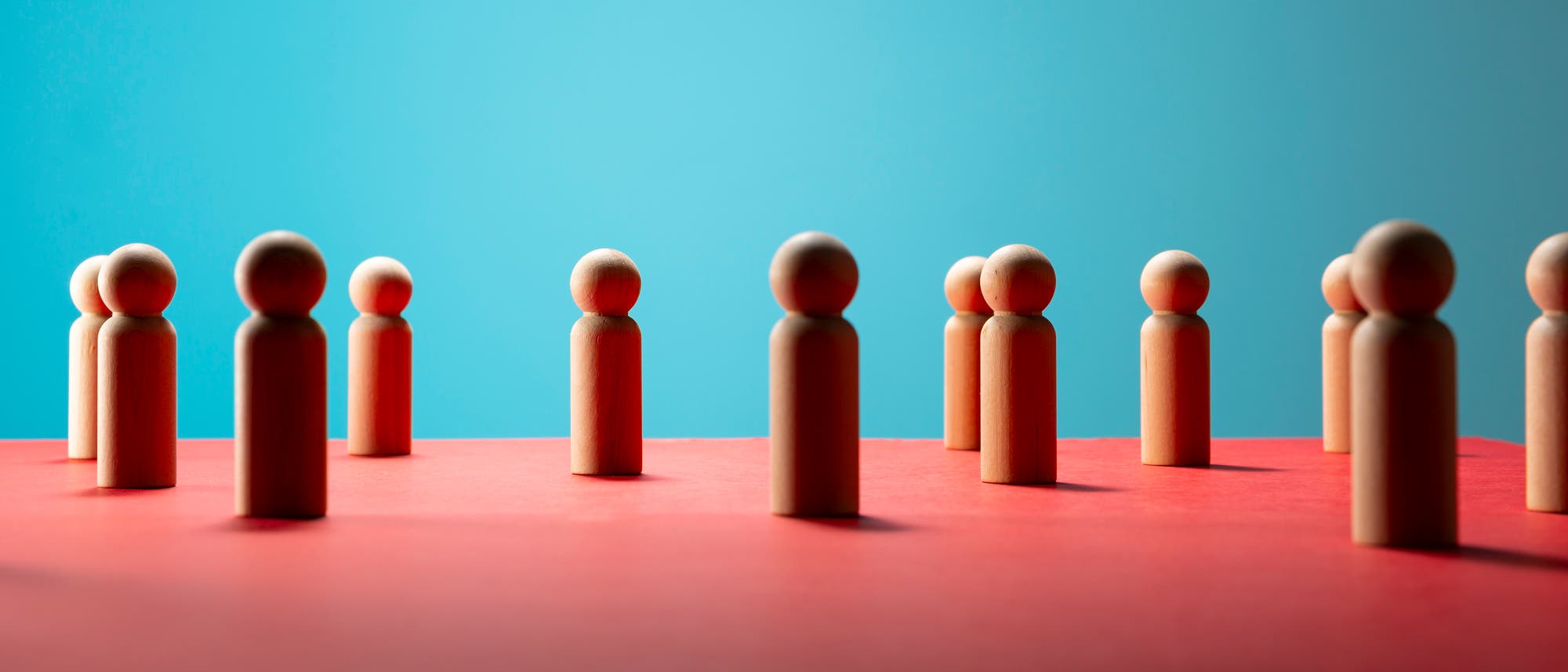 Holzfiguren stehen auf einer roten Oberfläche vor einem blauen Hintergrund. Die Figuren sind gleichmäßig verteilt und symbolisieren möglicherweise soziale Interaktion oder Teamarbeit. Die Szene ist minimalistisch und kontrastreich gestaltet. Holzfiguren stehen auf einer roten Oberfläche vor einem blauen Hintergrund. Die Figuren sind gleichmäßig verteilt und symbolisieren möglicherweise soziale Interaktion oder Teamarbeit. Die Szene ist minimalistisch und kontrastreich gestaltet.