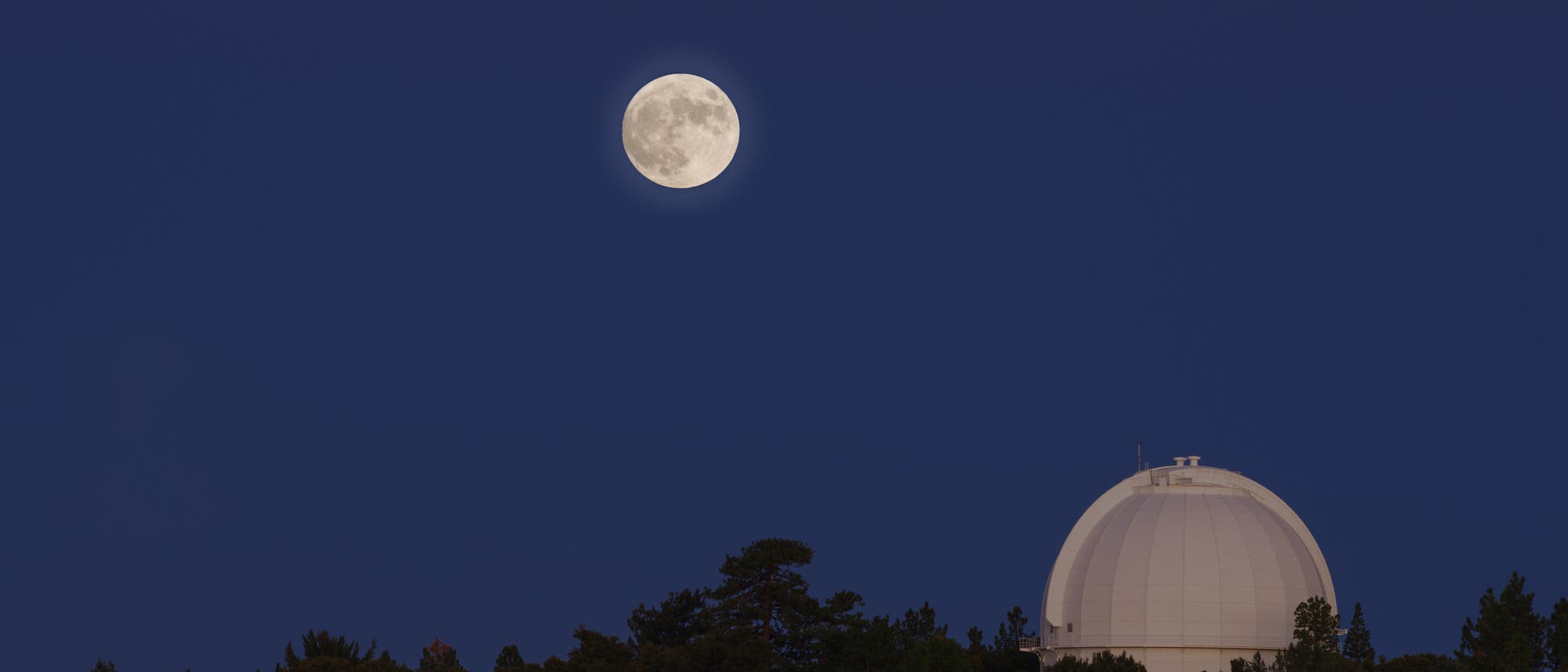 Ein fast voller Mond leuchtet am klaren Nachthimmel &uuml;ber einem Observatorium, das von B&auml;umen umgeben ist. Die Kuppel des Observatoriums ist gut sichtbar und hebt sich gegen den dunklen Himmel ab. Es gibt keine Menschen auf dem Bild.