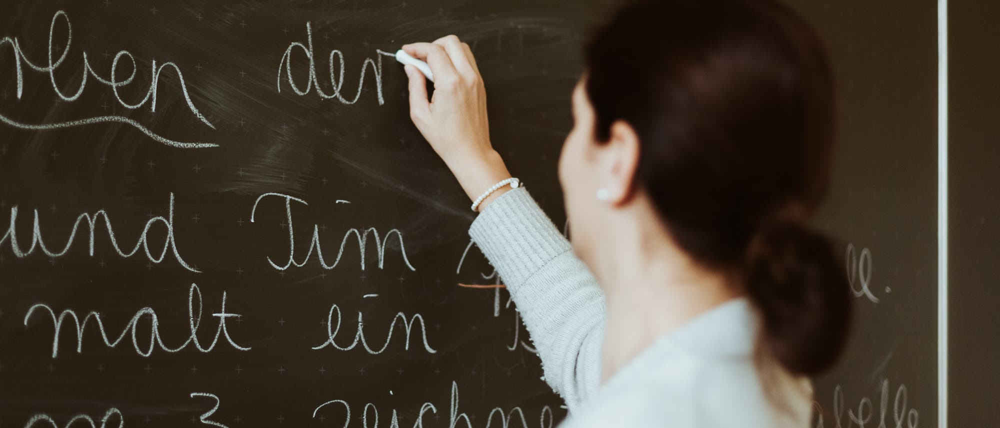 Eine Person schreibt mit Kreide auf eine dunkle Tafel. Der Text auf der Tafel ist in deutscher Schreibschrift verfasst und enth&auml;lt W&ouml;rter wie "Tim" und "malt". Die Person tr&auml;gt einen hellen Pullover und ein Armband. Die Szene vermittelt eine Unterrichtssituation in einem Klassenzimmer.