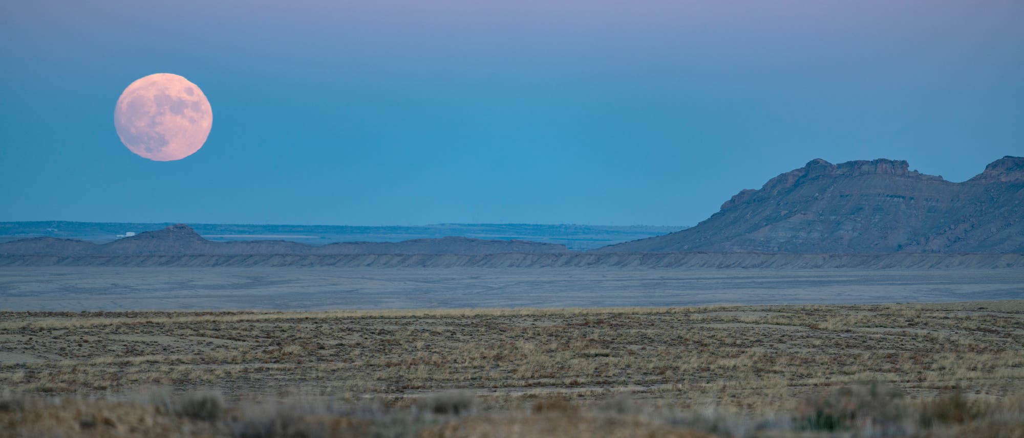 Eine weite, trockene Landschaft in New Mexico, USA bei Dämmerung mit einem großen, hellen Vollmond am Horizont. Der Himmel ist in sanften Blau- und Rosatönen gefärbt. 