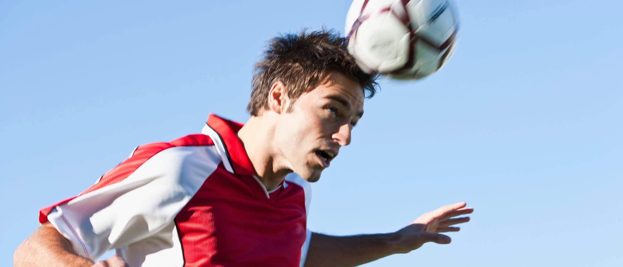 Ein Fußballspieler in einem rot-weißen Trikot köpft einen Ball. Der Himmel ist klar und blau. Der Spieler zeigt Konzentration und Bewegung, während er den Ball mit dem Kopf trifft. Ein Fußballspieler in einem rot-weißen Trikot köpft einen Ball. Der Himmel ist klar und blau. Der Spieler zeigt Konzentration und Bewegung, während er den Ball mit dem Kopf trifft.