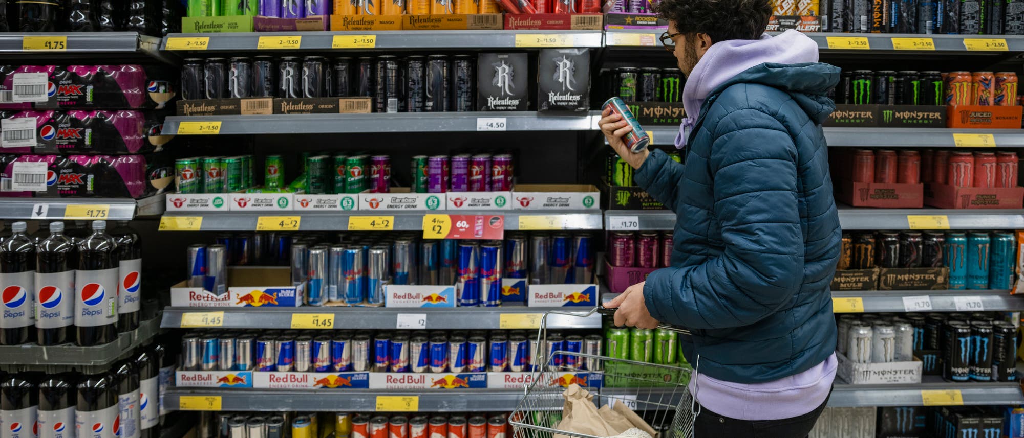 Eine Person mit Brille und lockigem Haar steht in einem Supermarkt vor einem Regal mit verschiedenen Energydrinks und Softdrinks, darunter Marken wie Red Bull, Monster und Pepsi. Sie trägt eine blaue Jacke und hält in der linken Hand einen Einkaufskorb. Die Person scheint die Preise oder Informationen auf einer Getränkedose zu überprüfen. Die Regale sind mit Preisschildern versehen. Eine Person mit Brille und lockigem Haar steht in einem Supermarkt vor einem Regal mit verschiedenen Energydrinks und Softdrinks, darunter Marken wie Red Bull, Monster und Pepsi. Sie trägt eine blaue Jacke und hält in der linken Hand einen Einkaufskorb. Die Person scheint die Preise oder Informationen auf einer Getränkedose zu überprüfen. Die Regale sind mit Preisschildern versehen.