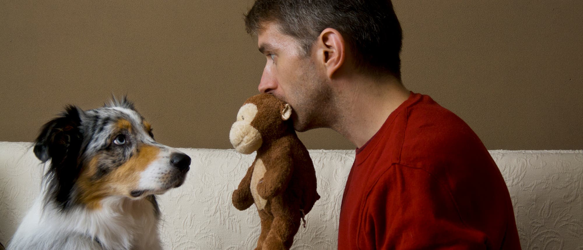 Ein Mann in einem roten Pullover h&auml;lt ein Pl&uuml;schtier in Form eines Affen im Mund und schaut einen Hund an, der aufmerksam zur&uuml;ckblickt. Mensch und Hund sitzen auf einem hellen Sofa vor einer dunklen Wand. Die Szene wirkt humorvoll und zeigt eine Interaktion zwischen Mensch und Tier.