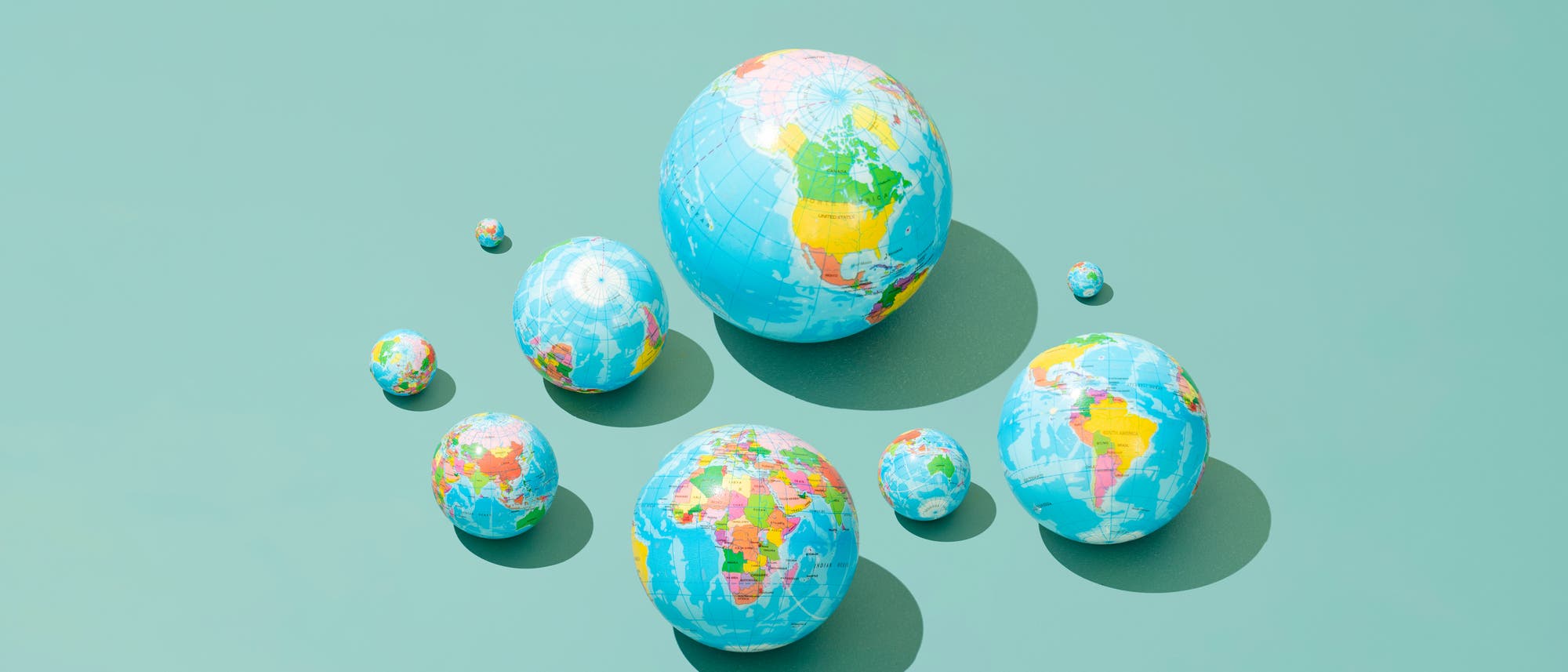 Eine Sammlung von neun unterschiedlich großen Weltkugeln, die auf einer hellgrünen Oberfläche angeordnet sind. Jede Kugel zeigt eine geografische Karte der Erde mit farblich hervorgehobenen Kontinenten und Ozeanen. Die Kugeln werfen Schatten auf die Oberfläche, was den Eindruck von Dreidimensionalität verstärkt. Die Anordnung der Kugeln vermittelt ein Gefühl von Vielfalt und Globalität. Eine Sammlung von neun unterschiedlich großen Weltkugeln, die auf einer hellgrünen Oberfläche angeordnet sind. Jede Kugel zeigt eine geografische Karte der Erde mit farblich hervorgehobenen Kontinenten und Ozeanen. Die Kugeln werfen Schatten auf die Oberfläche, was den Eindruck von Dreidimensionalität verstärkt. Die Anordnung der Kugeln vermittelt ein Gefühl von Vielfalt und Globalität.