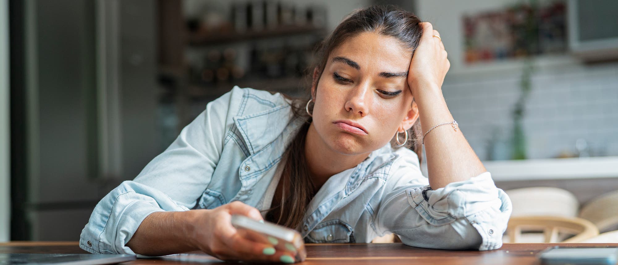 Eine Frau sitzt an einem Holztisch in einer Küche und schaut gelangweilt auf ihr Smartphone. Sie stützt ihren Kopf auf eine Hand. Im Hintergrund sind unscharf Küchenschränke und Pflanzen zu sehen. Die Szene vermittelt einen Eindruck von Langeweile oder Warten. Eine Frau sitzt an einem Holztisch in einer Küche und schaut gelangweilt auf ihr Smartphone. Sie stützt ihren Kopf auf eine Hand. Im Hintergrund sind unscharf Küchenschränke und Pflanzen zu sehen. Die Szene vermittelt einen Eindruck von Langeweile oder Warten.