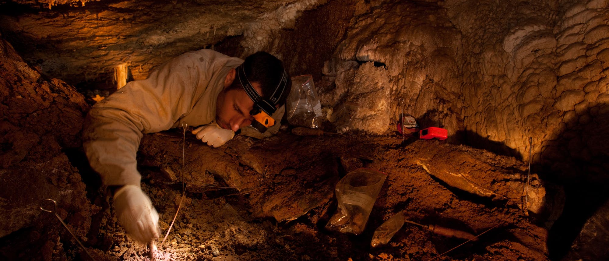 Eine Person mit Stirnlampe und Handschuhen untersucht den Boden einer Höhle. Die Umgebung ist dunkel und von erdigen, felsigen Strukturen geprägt. Im Vordergrund sind Werkzeuge und Probenbehälter sichtbar. Die Szene deutet auf eine archäologische oder geologische Untersuchung hin. Eine Person mit Stirnlampe und Handschuhen untersucht den Boden einer Höhle. Die Umgebung ist dunkel und von erdigen, felsigen Strukturen geprägt. Im Vordergrund sind Werkzeuge und Probenbehälter sichtbar. Die Szene deutet auf eine archäologische oder geologische Untersuchung hin.