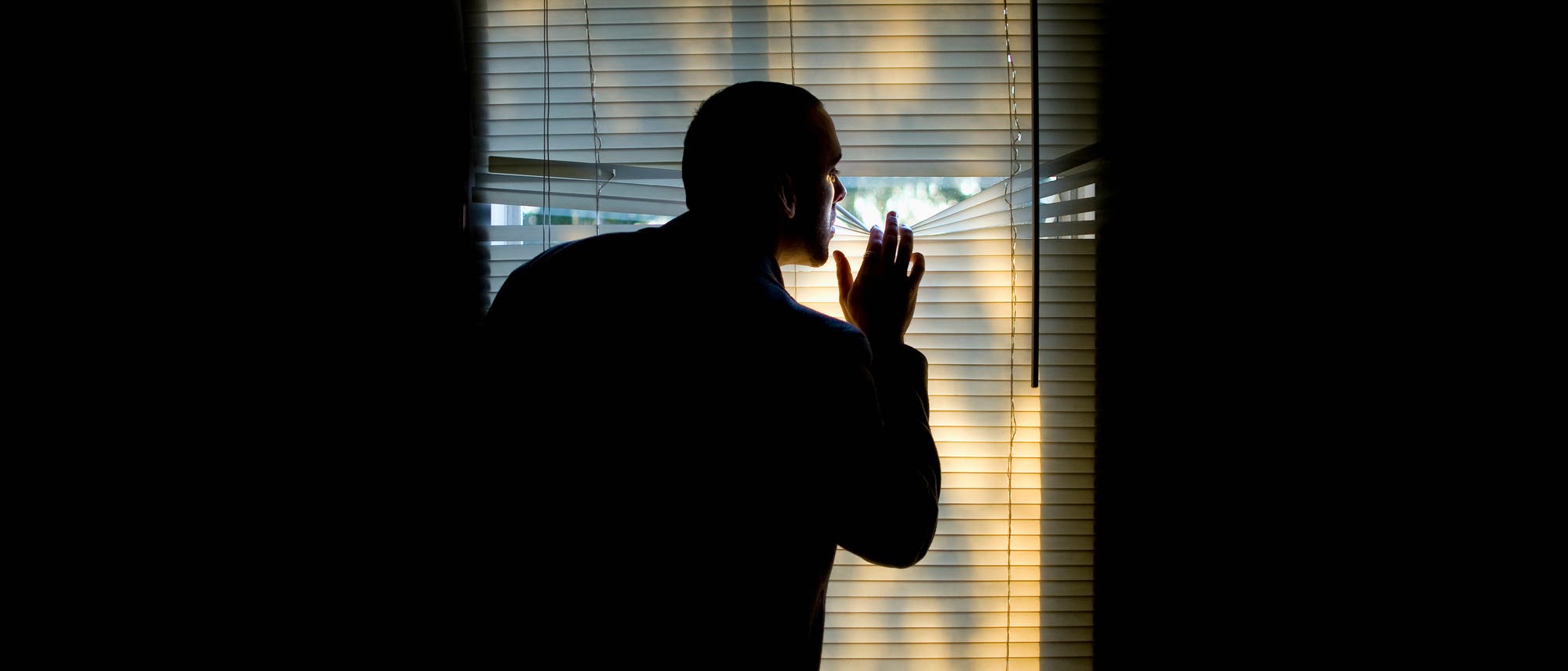 Eine Person steht in einem dunklen Raum und schaut durch die Jalousien eines Fensters nach drau&szlig;en. Die Szene ist von hinten beleuchtet, wodurch die Silhouette der Person hervorgehoben wird. Die Jalousien sind leicht ge&ouml;ffnet, sodass Lichtstrahlen in den Raum fallen.